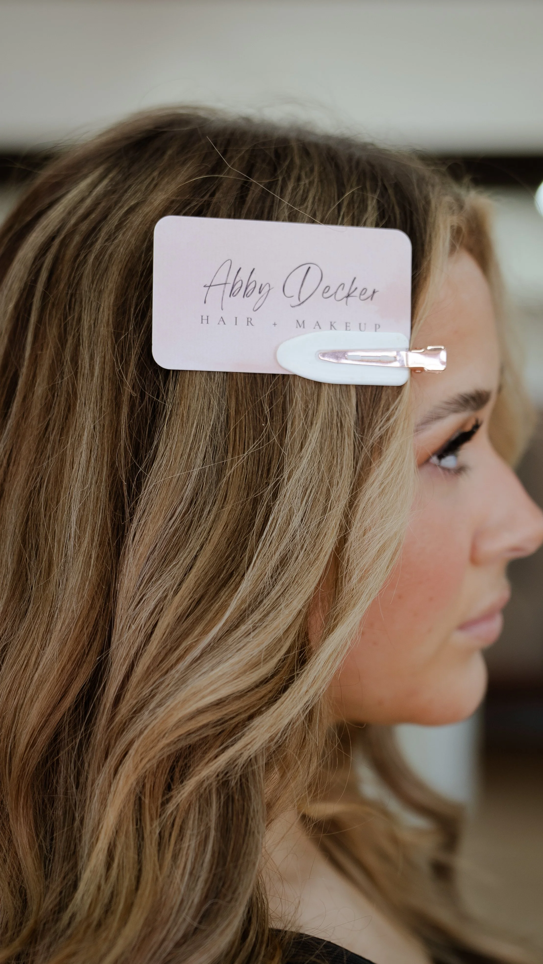 Close-up of a woman with long, wavy blonde hair, with a salon tag clipped to her hair that reads 'Abby Decker Hair + Makeup'.