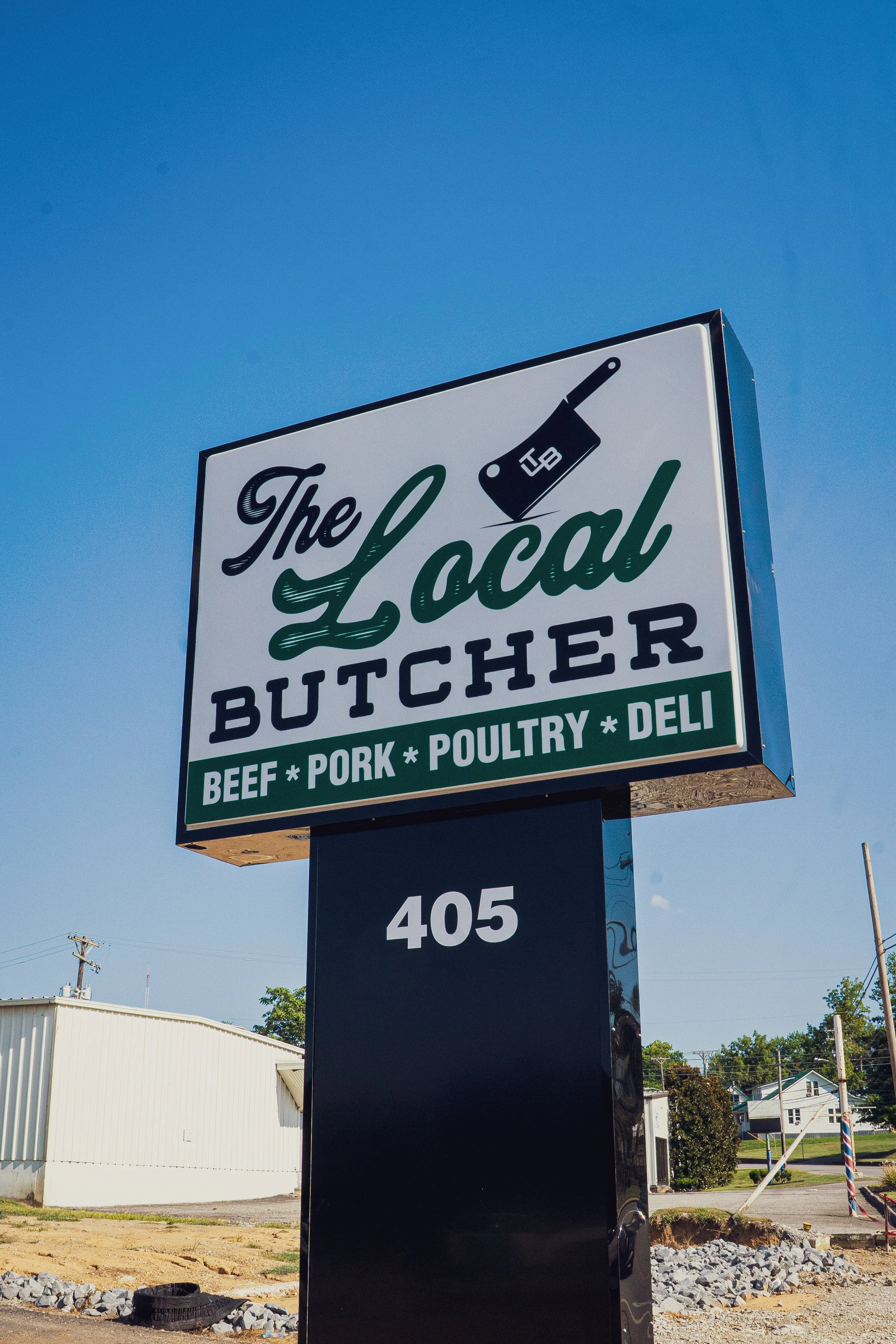 Sign for The Local Butcher advertising beef, pork, poultry, and deli, with the number 405 underneath, set against a clear blue sky.