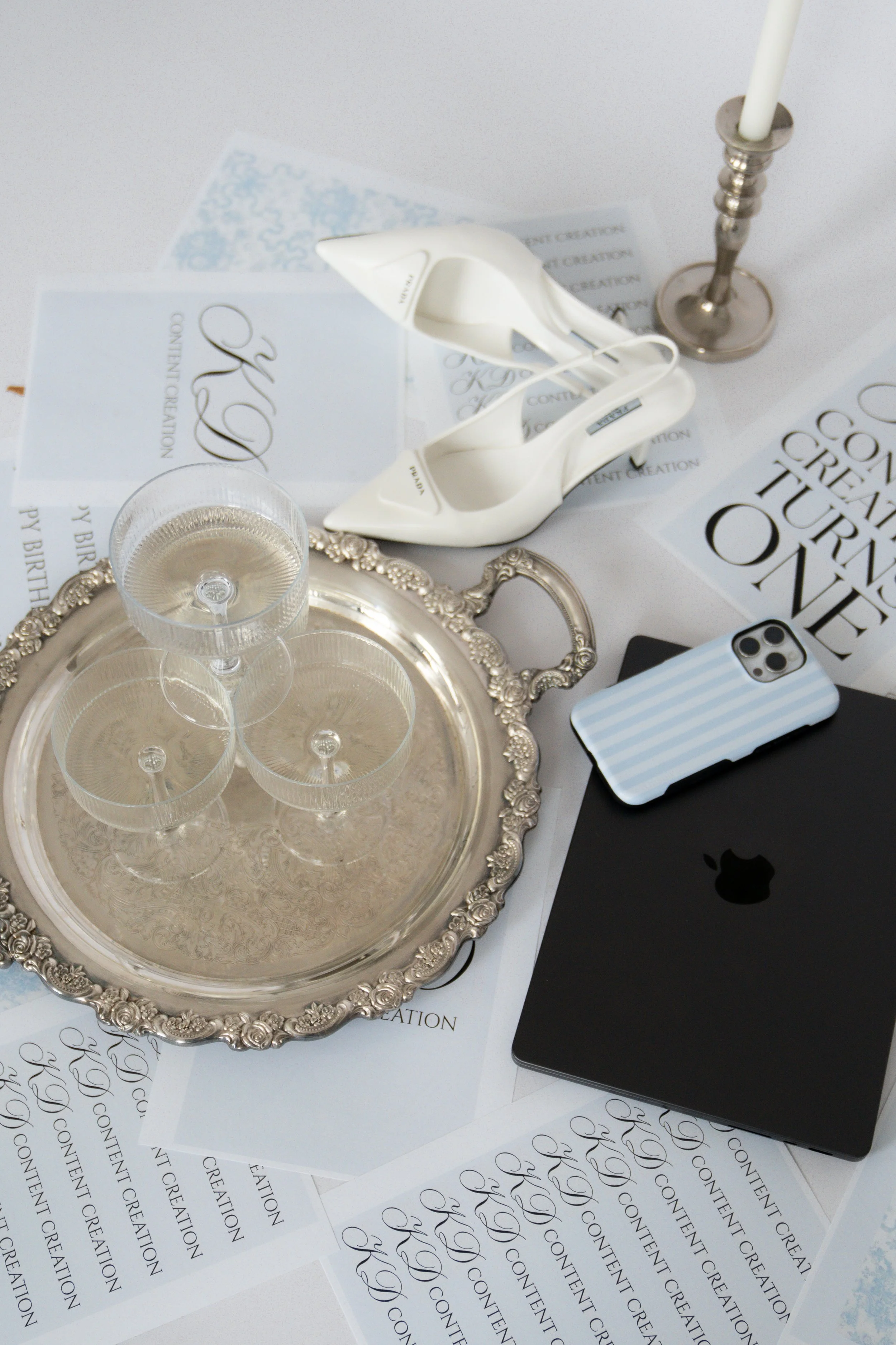 A flat lay of a table setting with a silver tray holding three cocktail glasses, white high-heeled shoes, a lit candle, a black phone with a striped case, and various printed papers on a white surface.