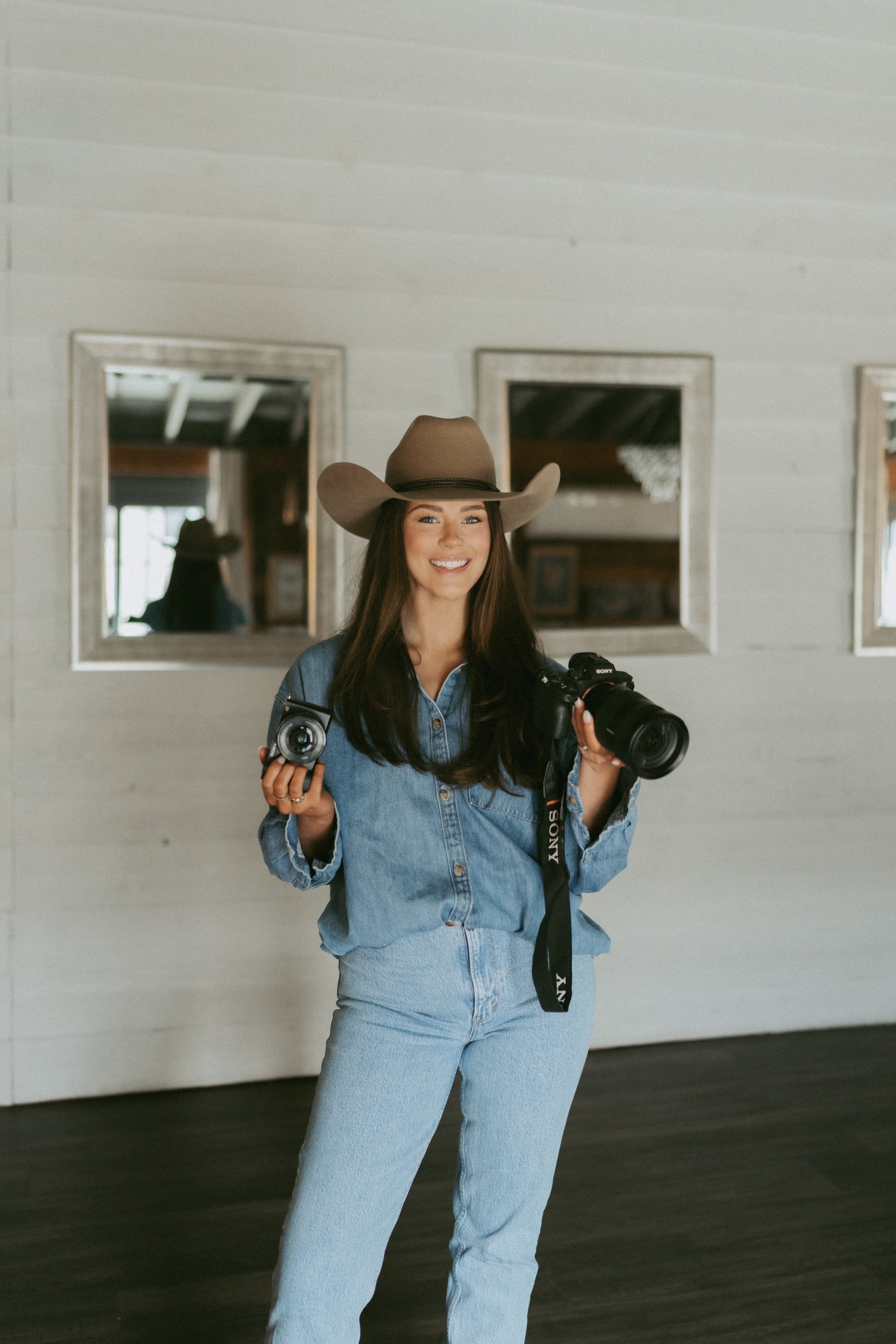 A young woman wearing a cowboy hat and denim outfit, holding a camera and a smaller device, smiling indoors with mirrors on the wall behind her.