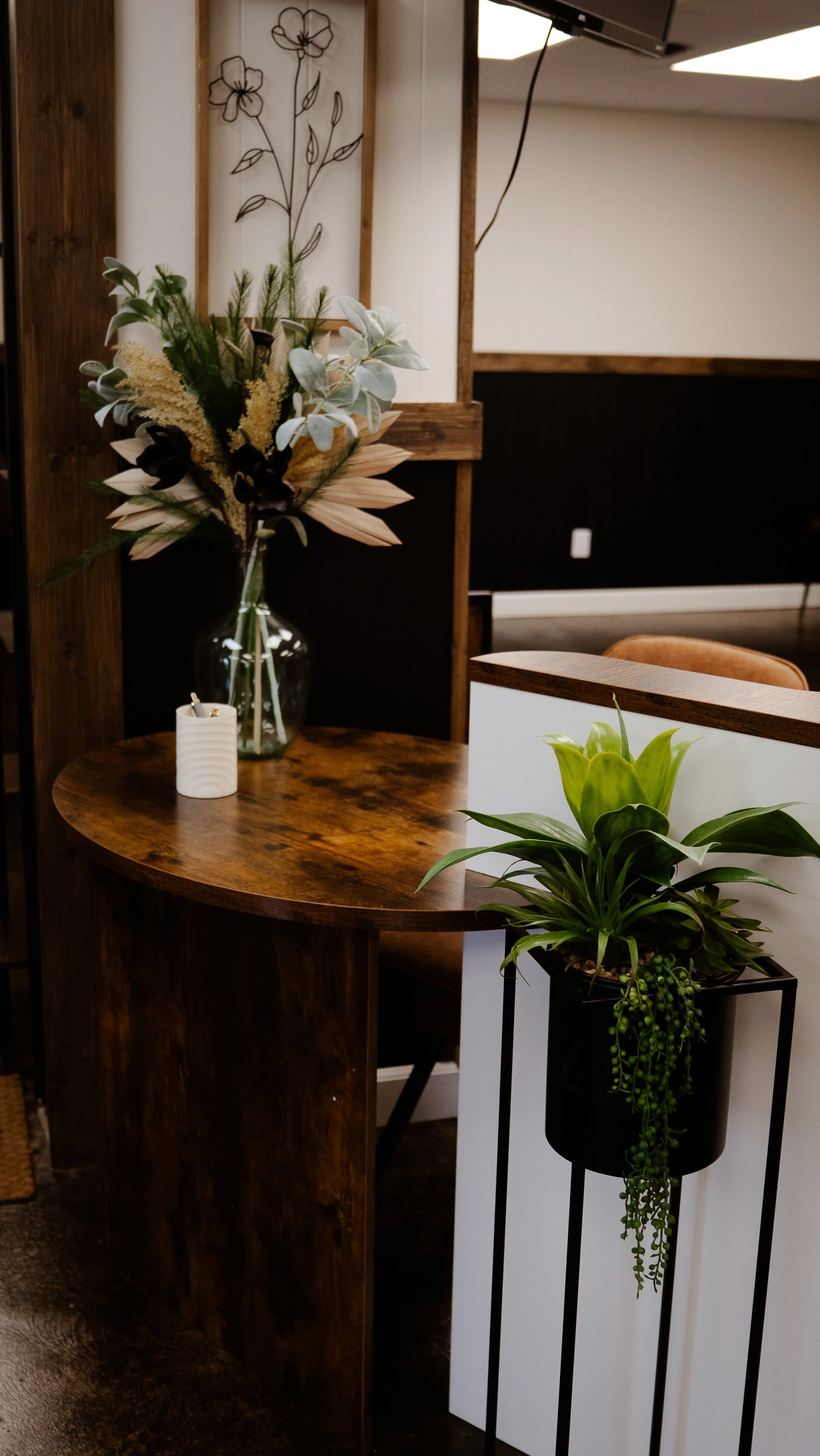 Indoor scene with a wooden table, a glass vase with dried flowers and greenery, a white candle, and a potted green plant on a black stand.
