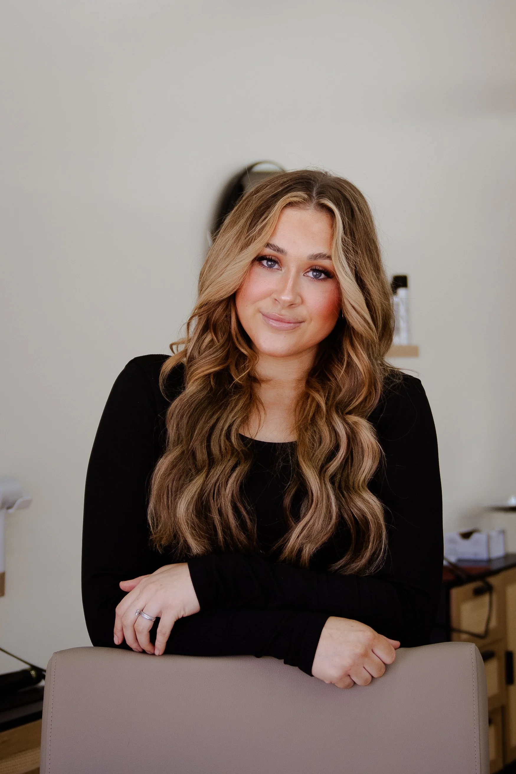 A young woman with long, wavy blonde hair and blue eyes, wearing a black long-sleeve top, is sitting at a table with her arms crossed on the back of a chair, smiling softly in an indoor setting.