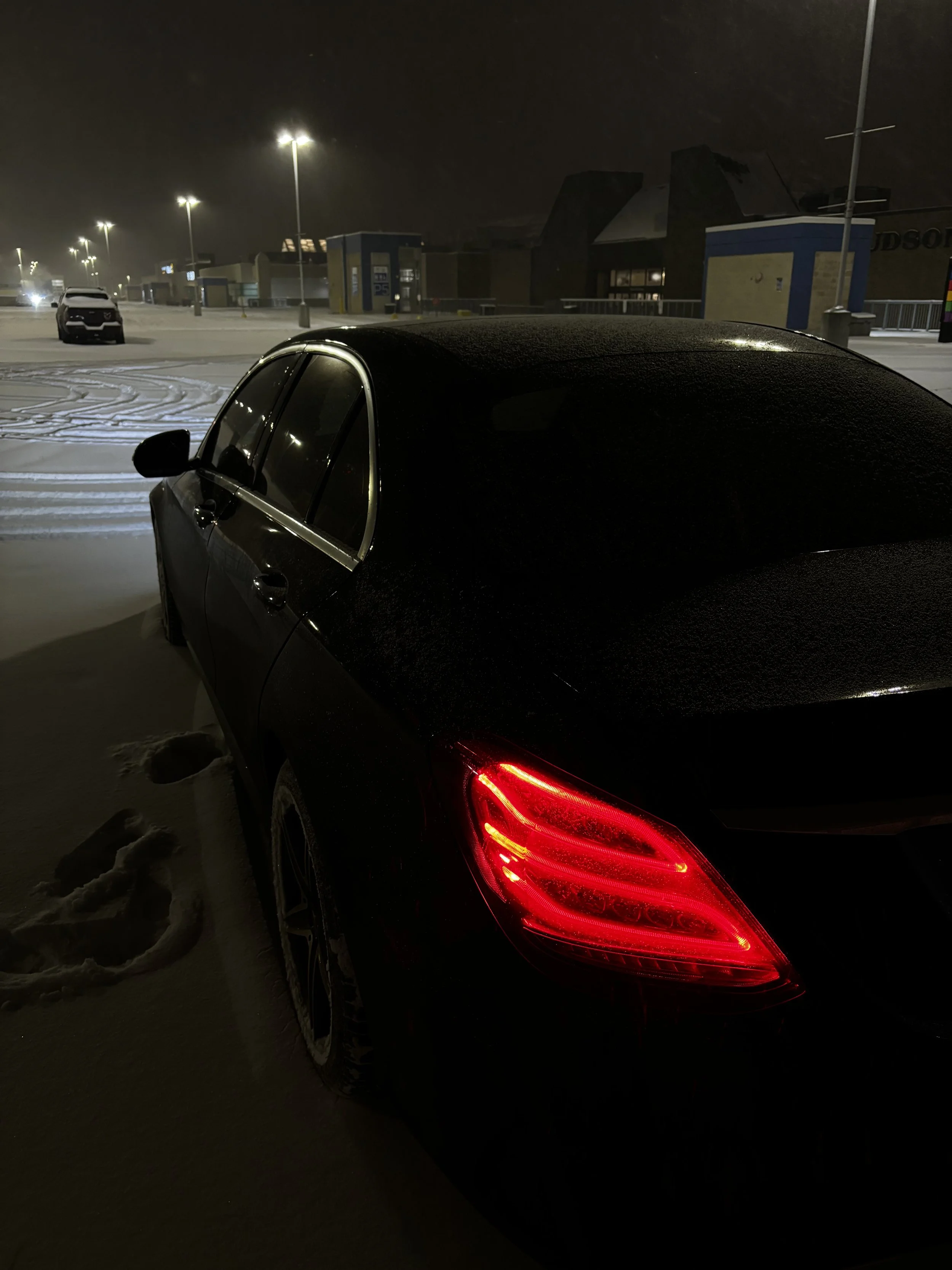 A black sedan parked in a snowy parking lot at night, with red taillights illuminated and snow-covered ground.