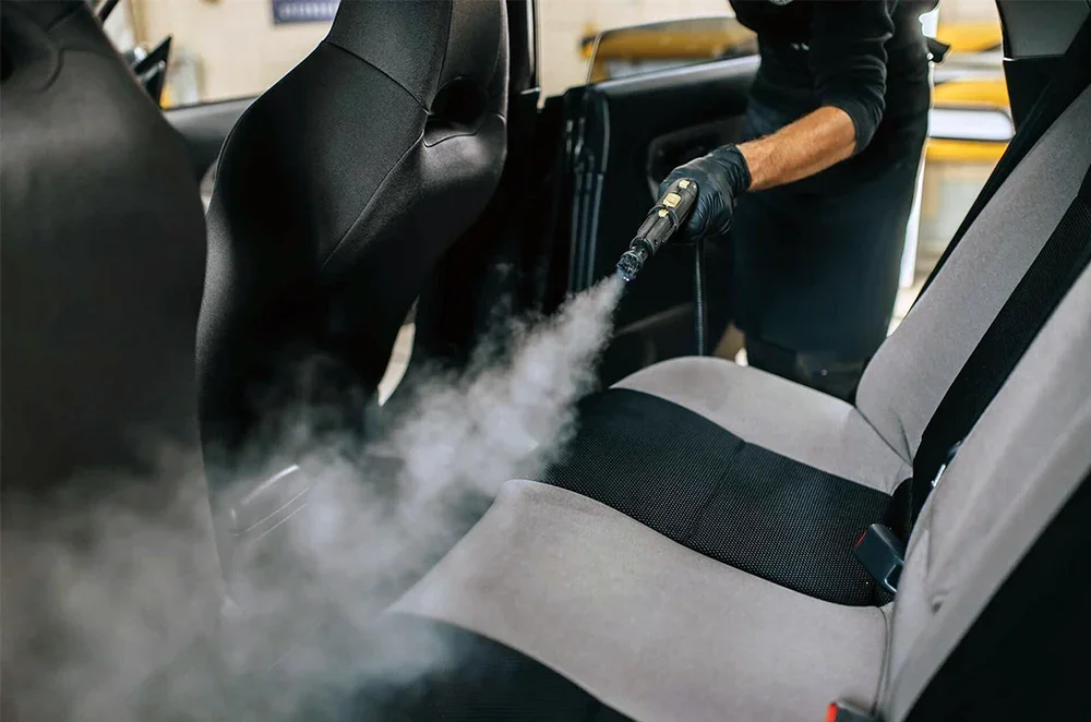 Person wearing black gloves using a steam cleaner on the back seat of a car, emitting steam to clean the upholstery.