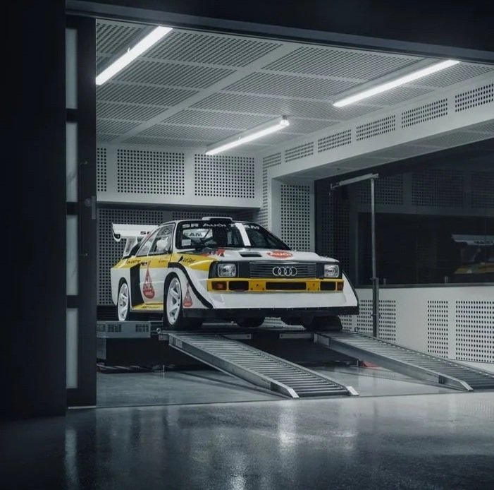 A white Audi Quattro rally car with yellow and black livery on a platform in a testing or storage facility, inside a modern room with perforated ceiling panels and bright overhead lighting.