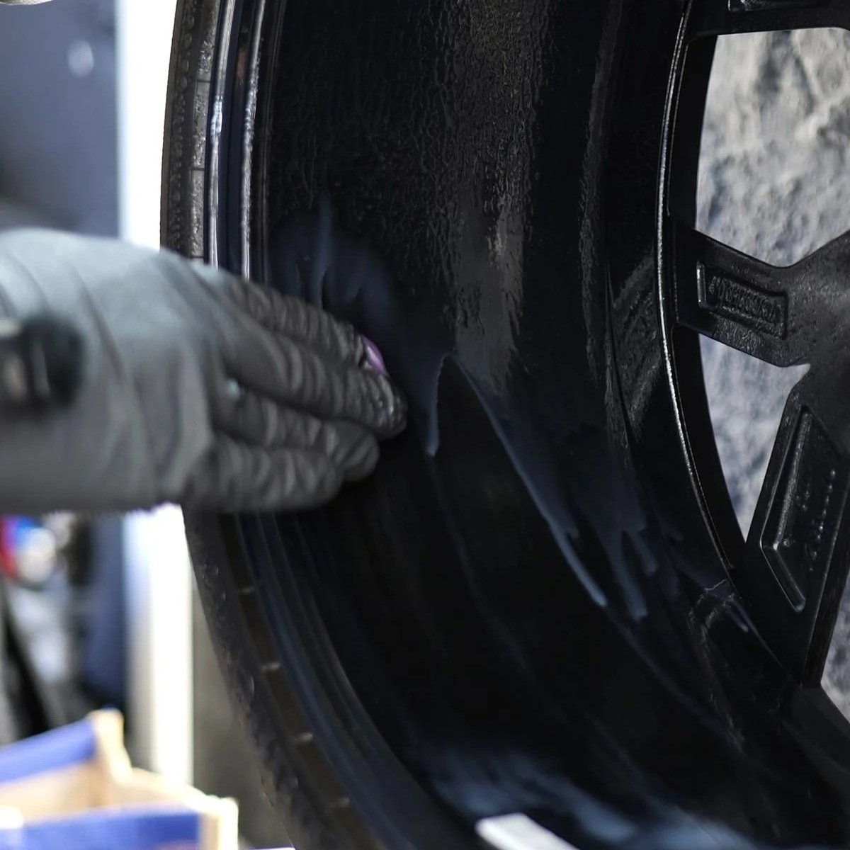 A person wearing a gray glove cleaning the inside of a black car wheel with a cloth.