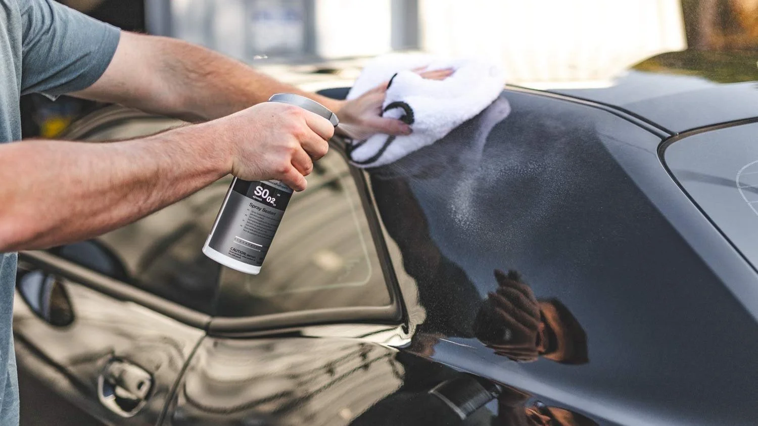 Person applying spray sealant to a black car's hood with a cloth.