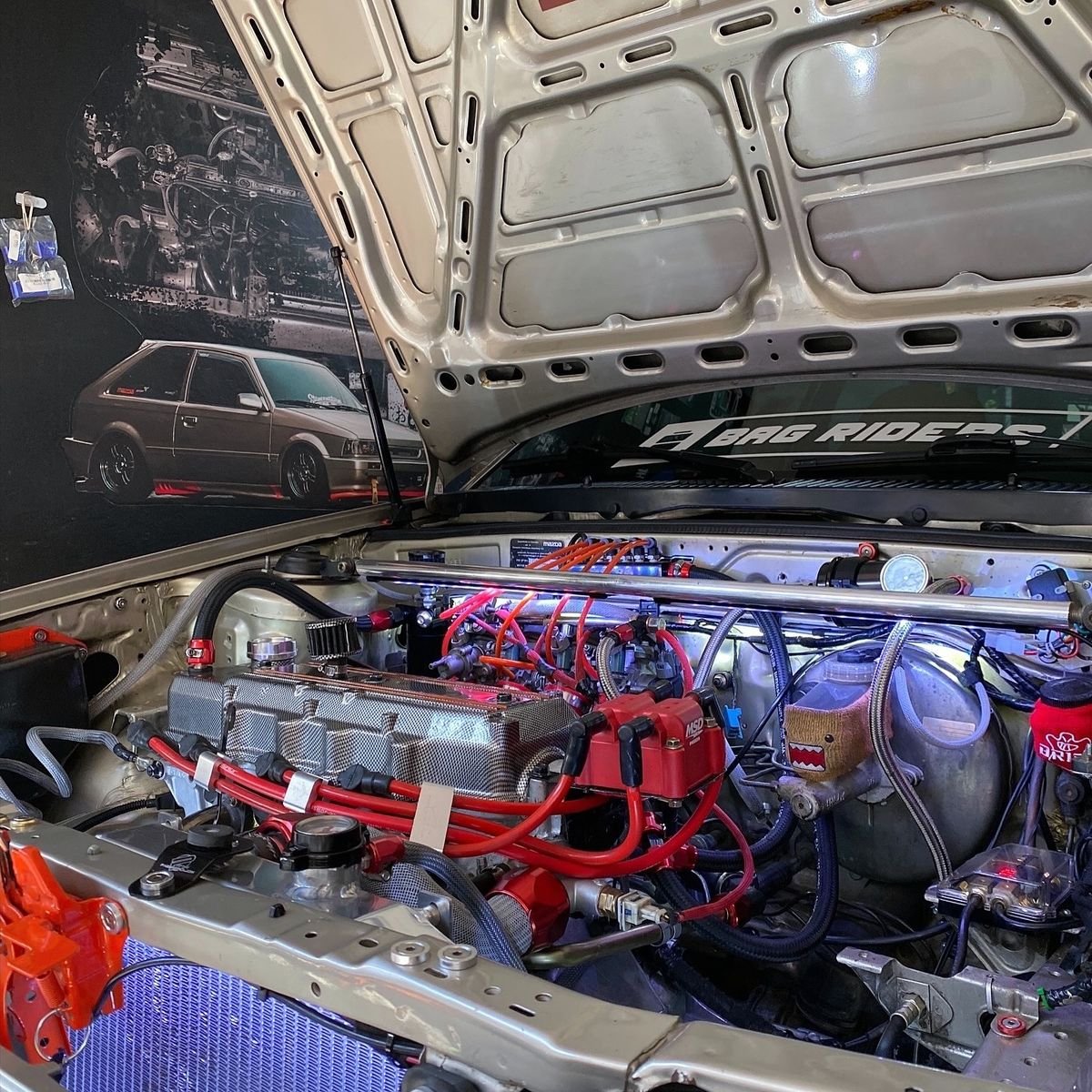 Inside view of a modified car engine with various colorful parts, wires, and components, with a poster of a classic hatchback car and a 'BAG RIDERS' sticker in the background.