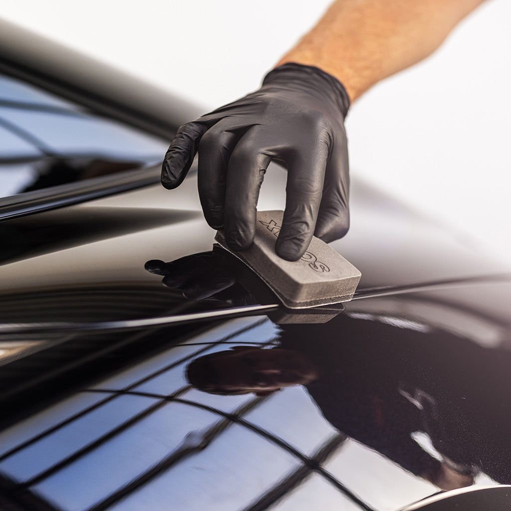 Person wearing black gloves polishing a black car with a sponge.