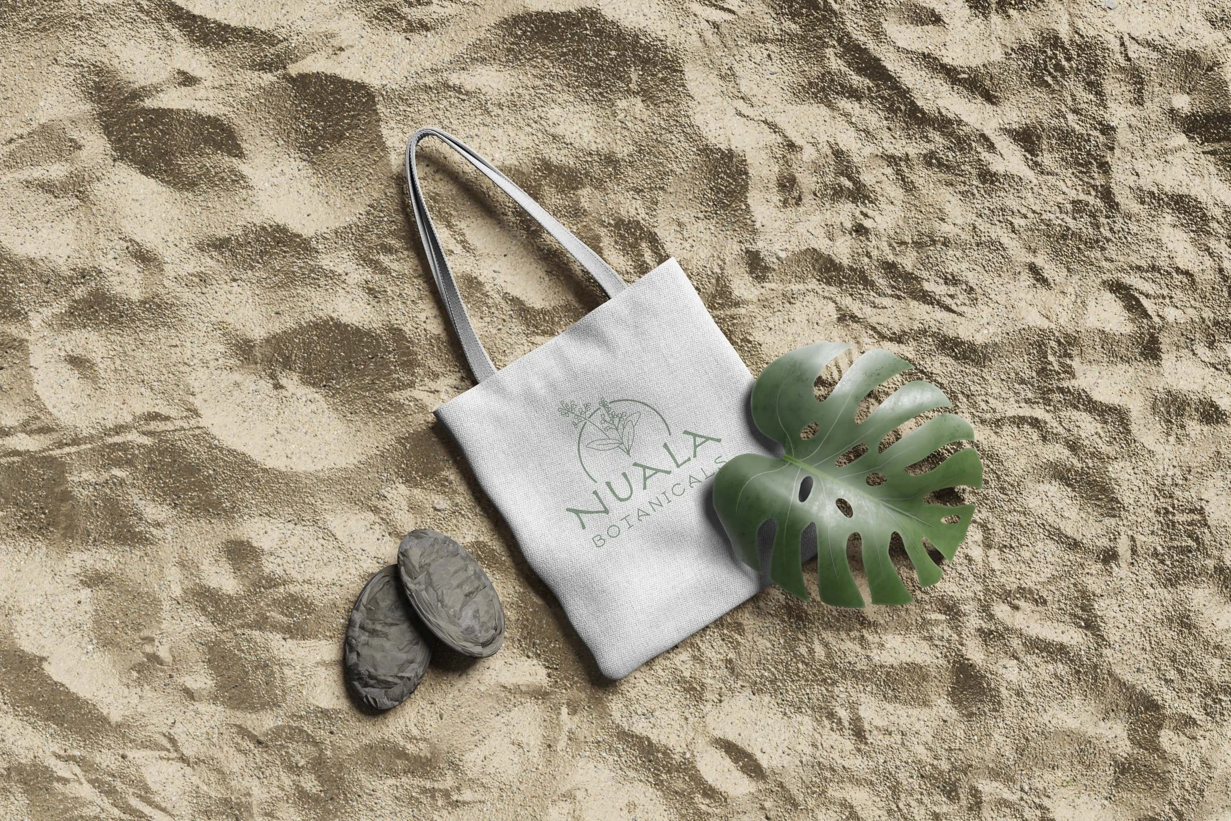 A tote bag labeled "NULAS BOTANICA" on sandy ground, with a green monstera leaf and two flat stones nearby.