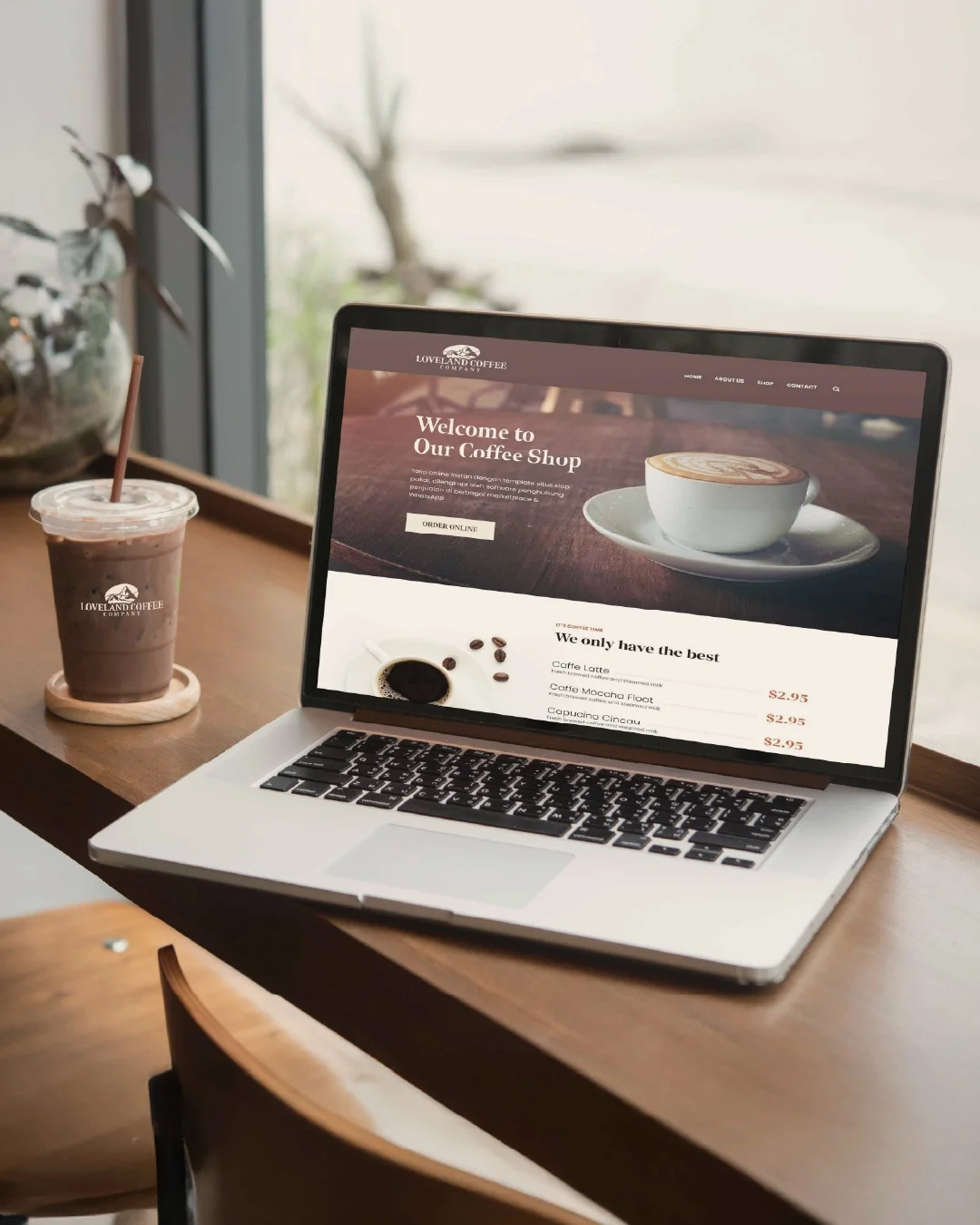 Laptop on wooden table displaying a coffee shop website, next to iced coffee drink in a transparent cup with a straw, in a cozy cafe setting with a window in the background.