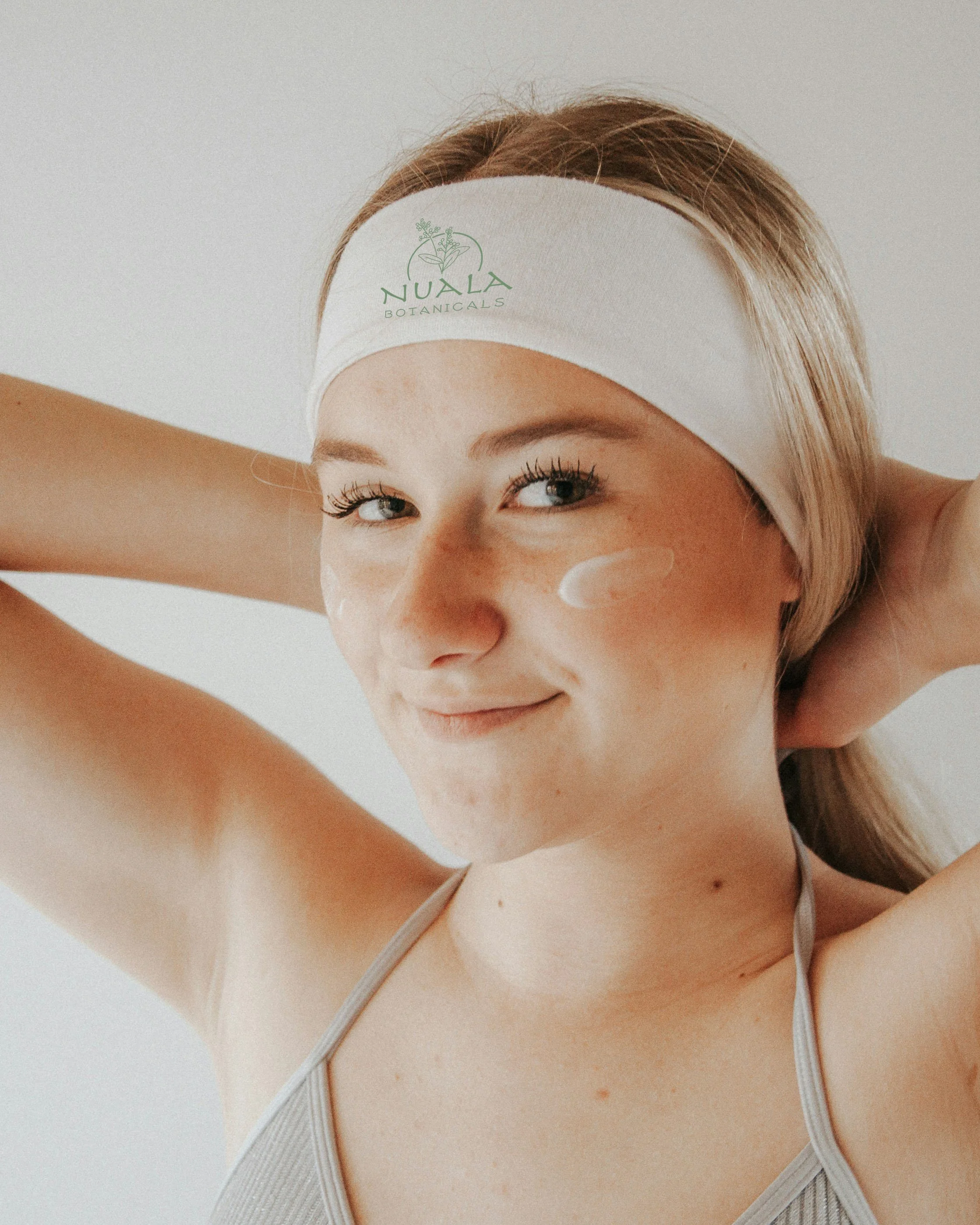 A young woman with blonde hair wearing a white headband that says 'Nuala Botanicals,' smiling and applying skincare cream to her cheek.