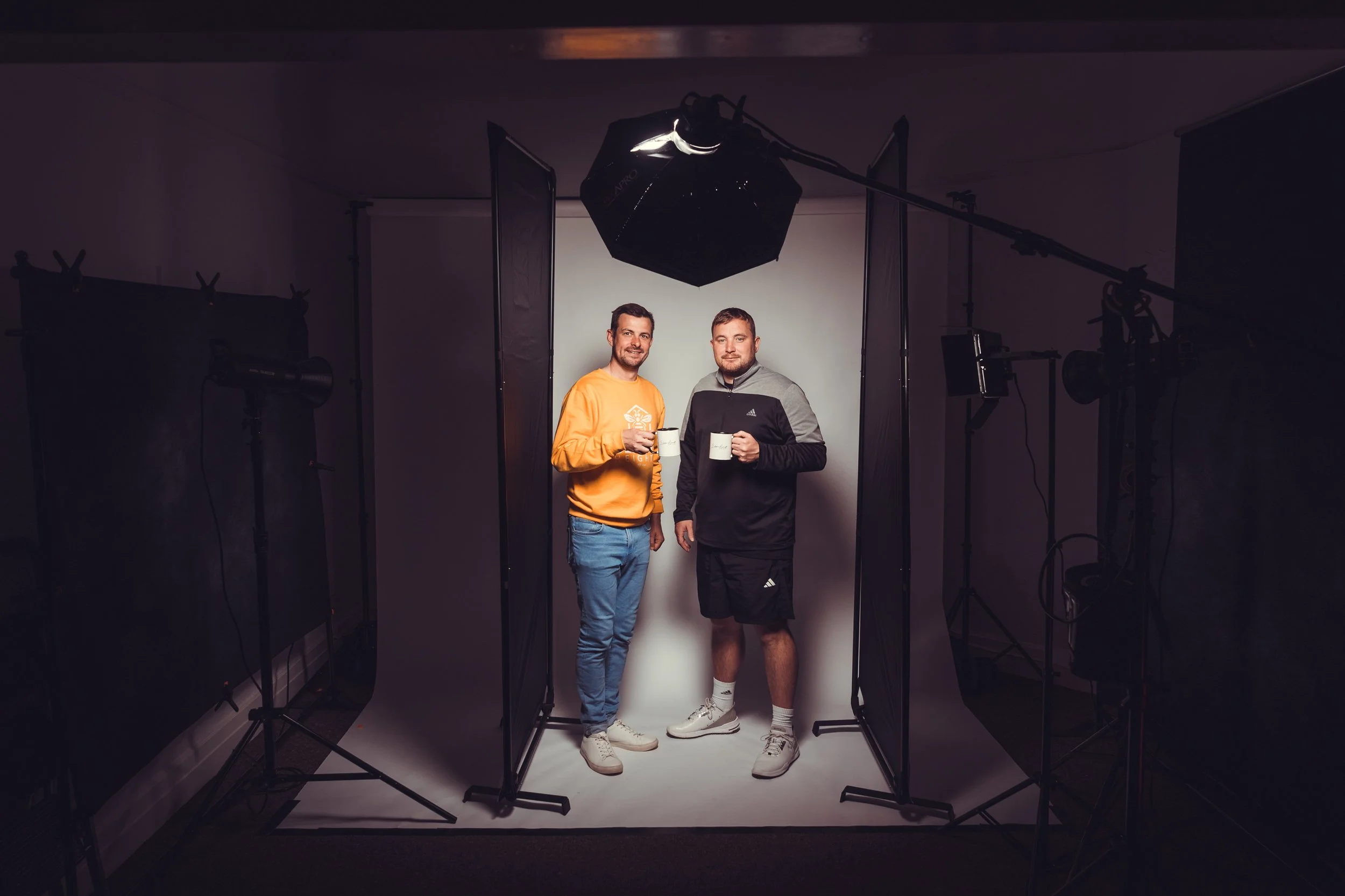 Two men standing in a photography studio, holding white mugs, surrounded by professional lighting and backdrop equipment.