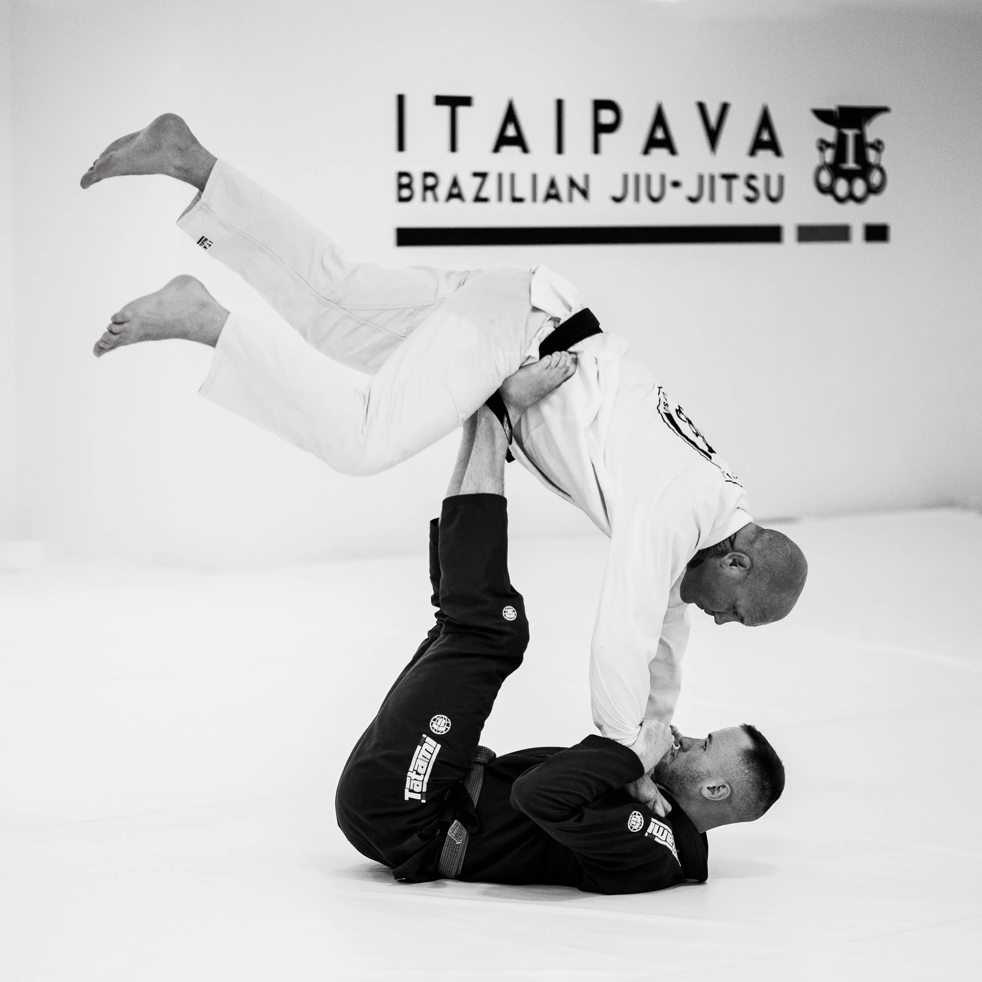 Two men practicing Brazilian Jiu-Jitsu on a mat; one man in a white gi is performing a submission move on a man in a black gi; background features a sign for ITAIPAVA Brazilian Jiu-Jitsu.