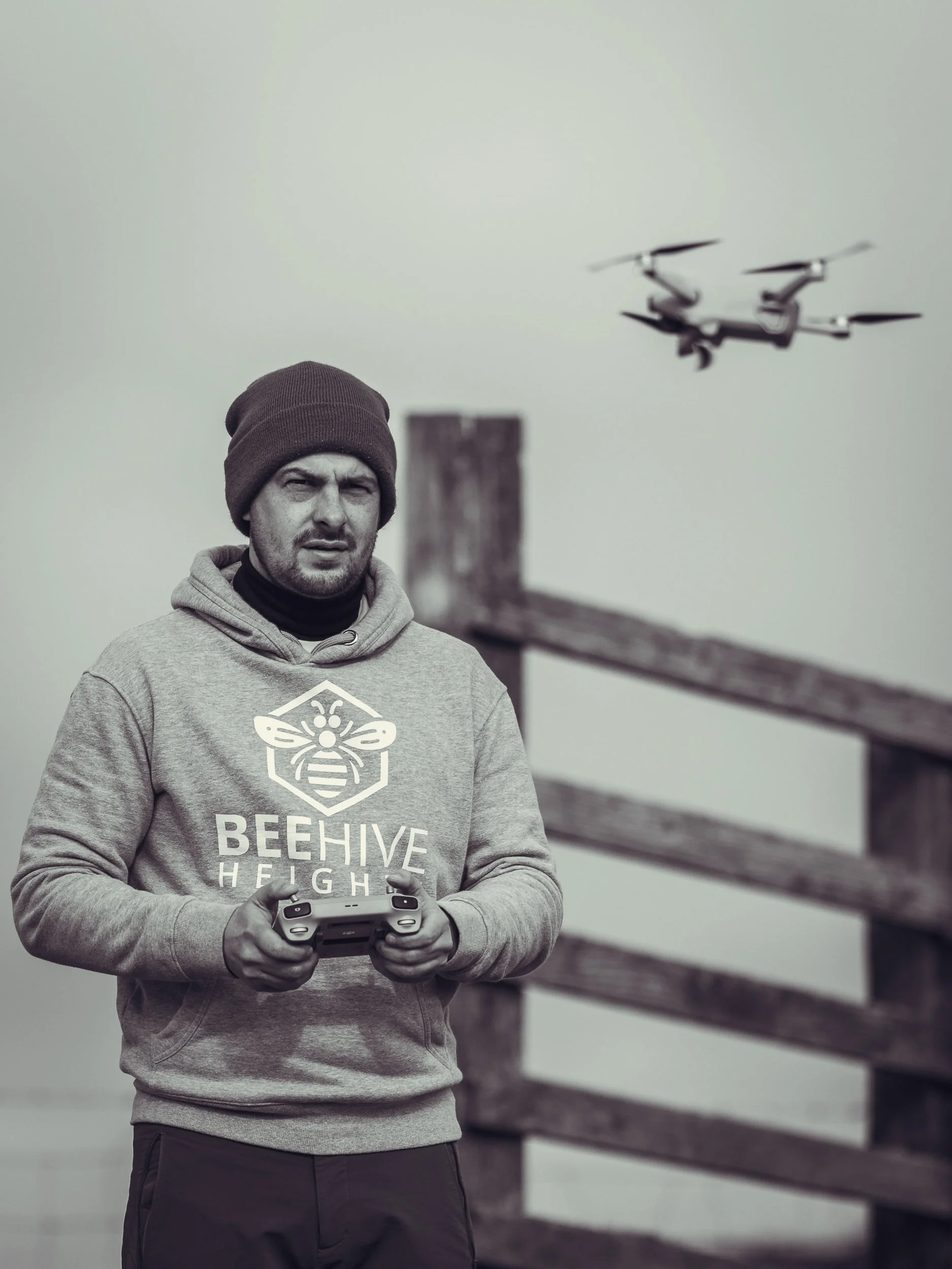 A man wearing a hoodie with a bee logo and the words "BEEHIVE HEIGHTS." He is holding a drone remote controller, standing outdoors near a wooden fence, with a drone flying in the background.
