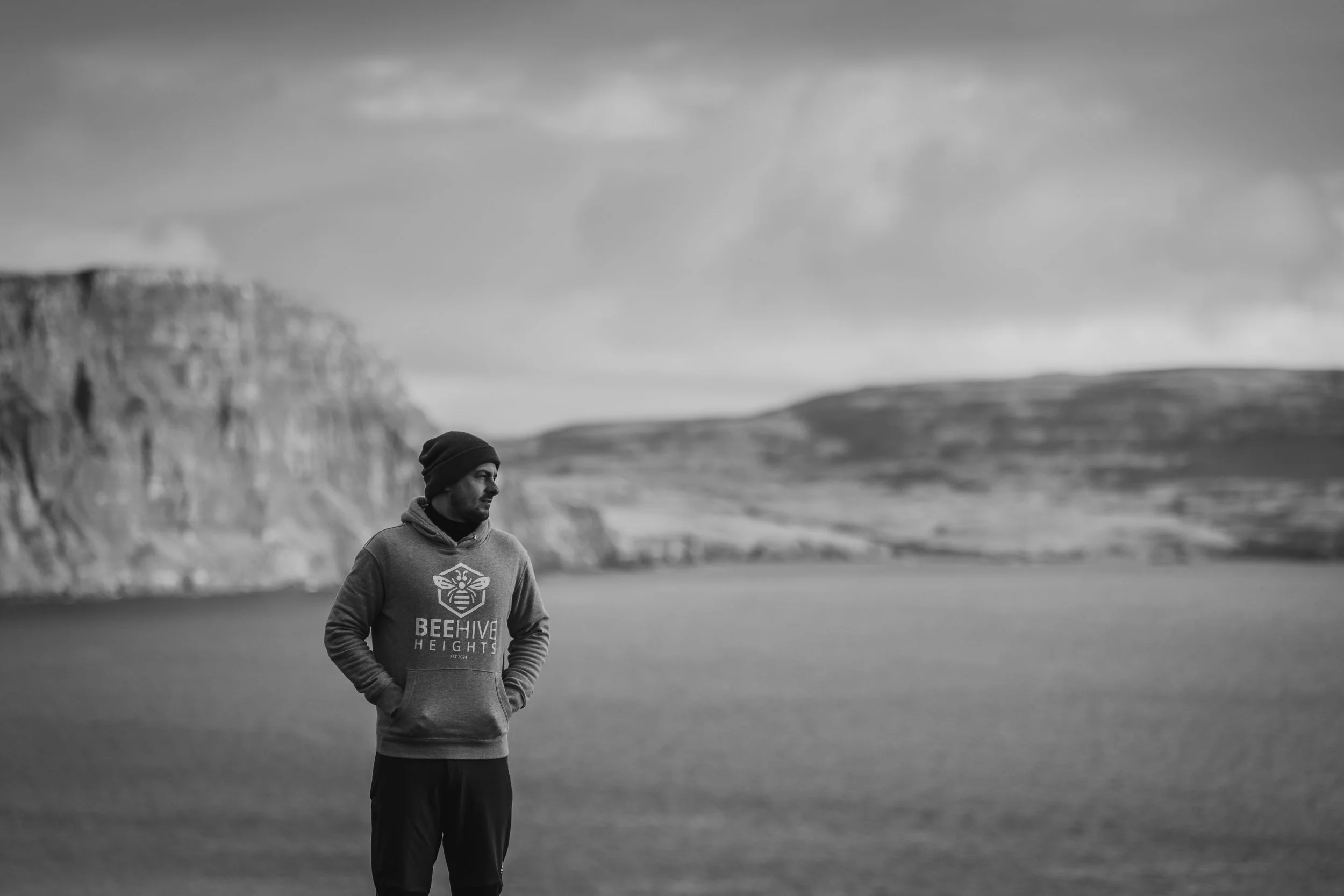 A man wearing a hoodie and beanie stands near a lake with mountains or cliffs in the background, looking to the side in a black and white photo.