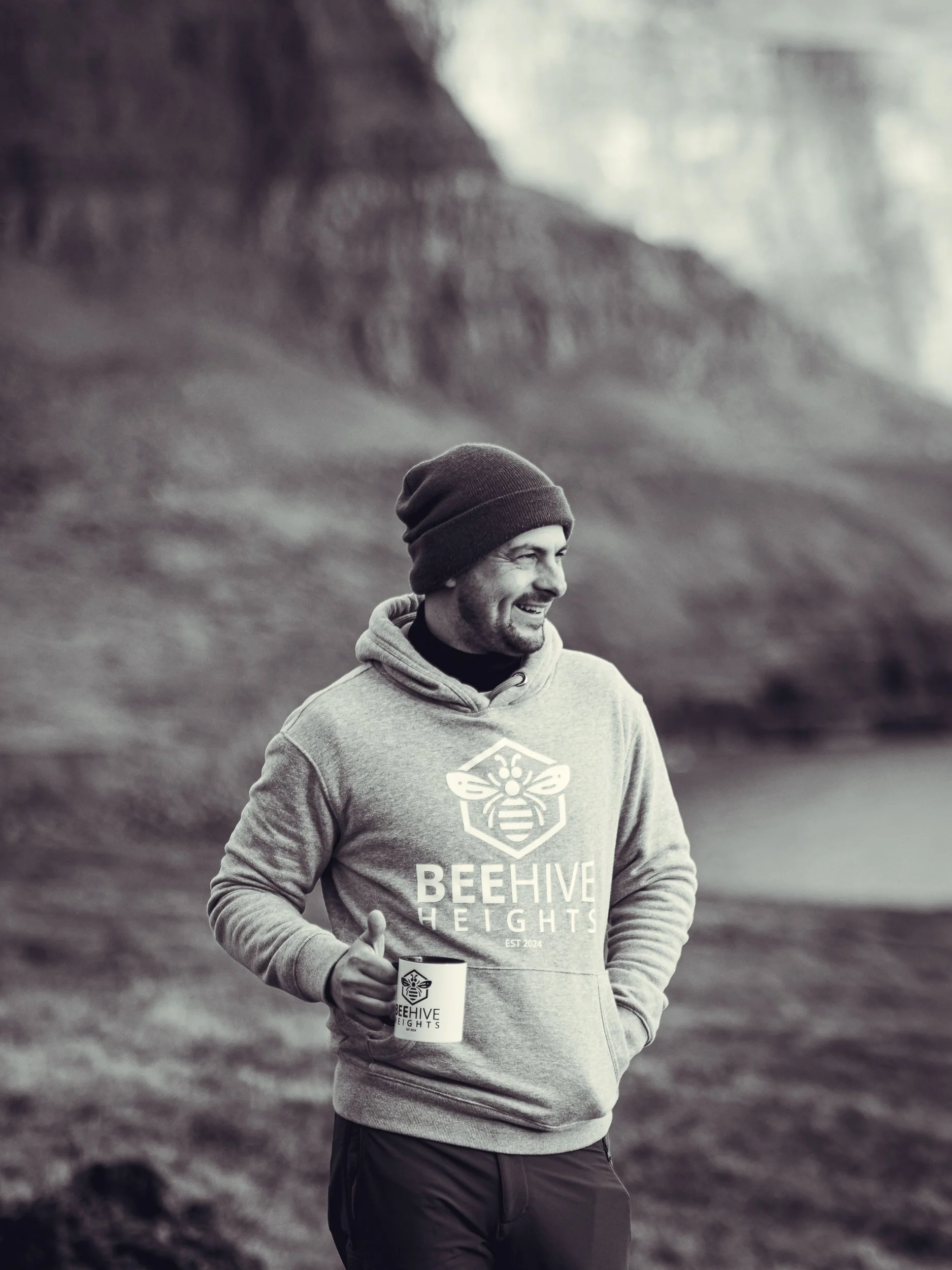 A man smiling and wearing a beanie and hoodie with the logo 'Beehive Heights' while holding a mug with the same logo outdoors near a rocky landscape.
