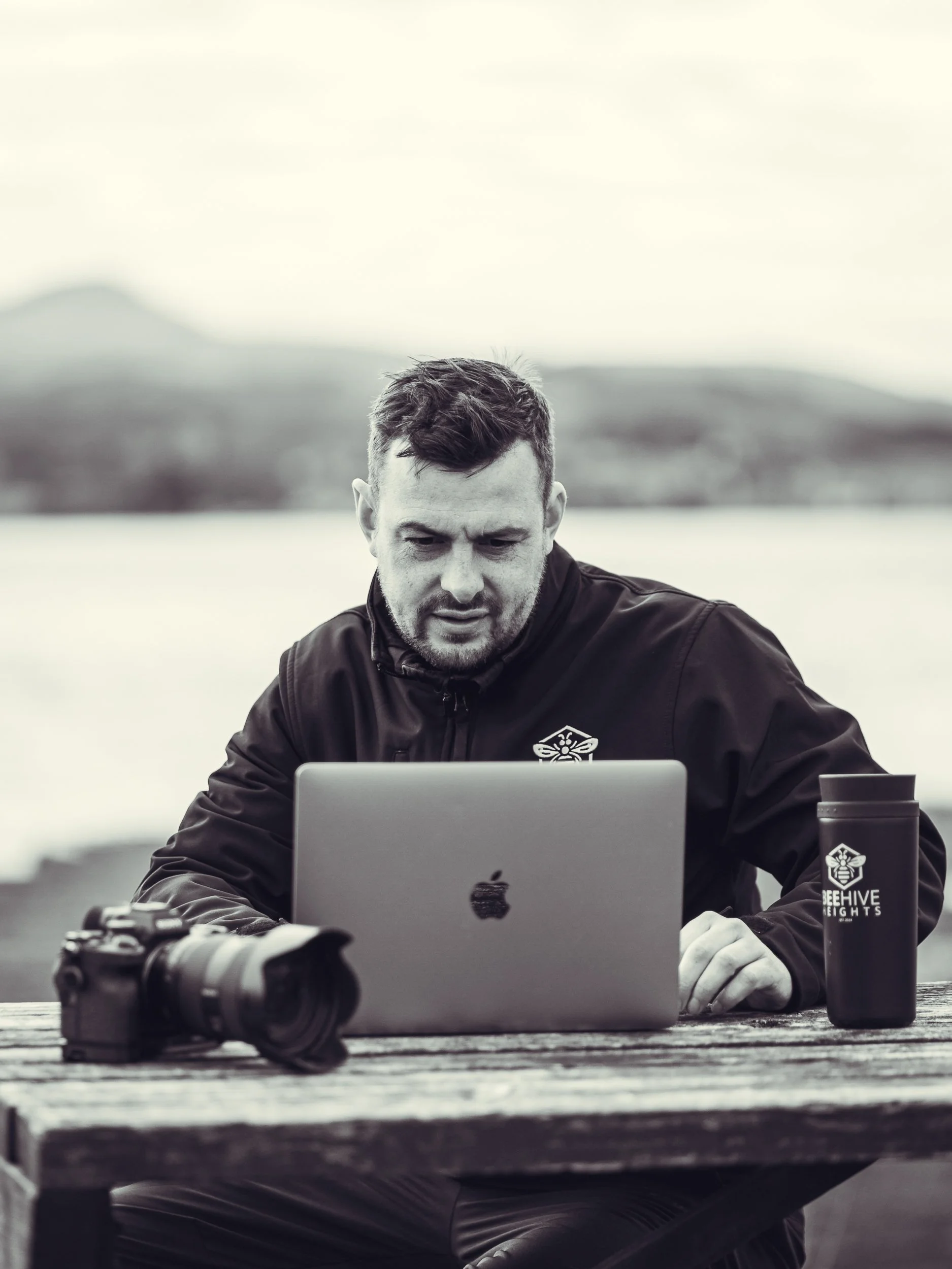 Man sitting at a wooden table outdoors using a MacBook with a camera and a travel mug nearby, overlooking a lake and distant hills.