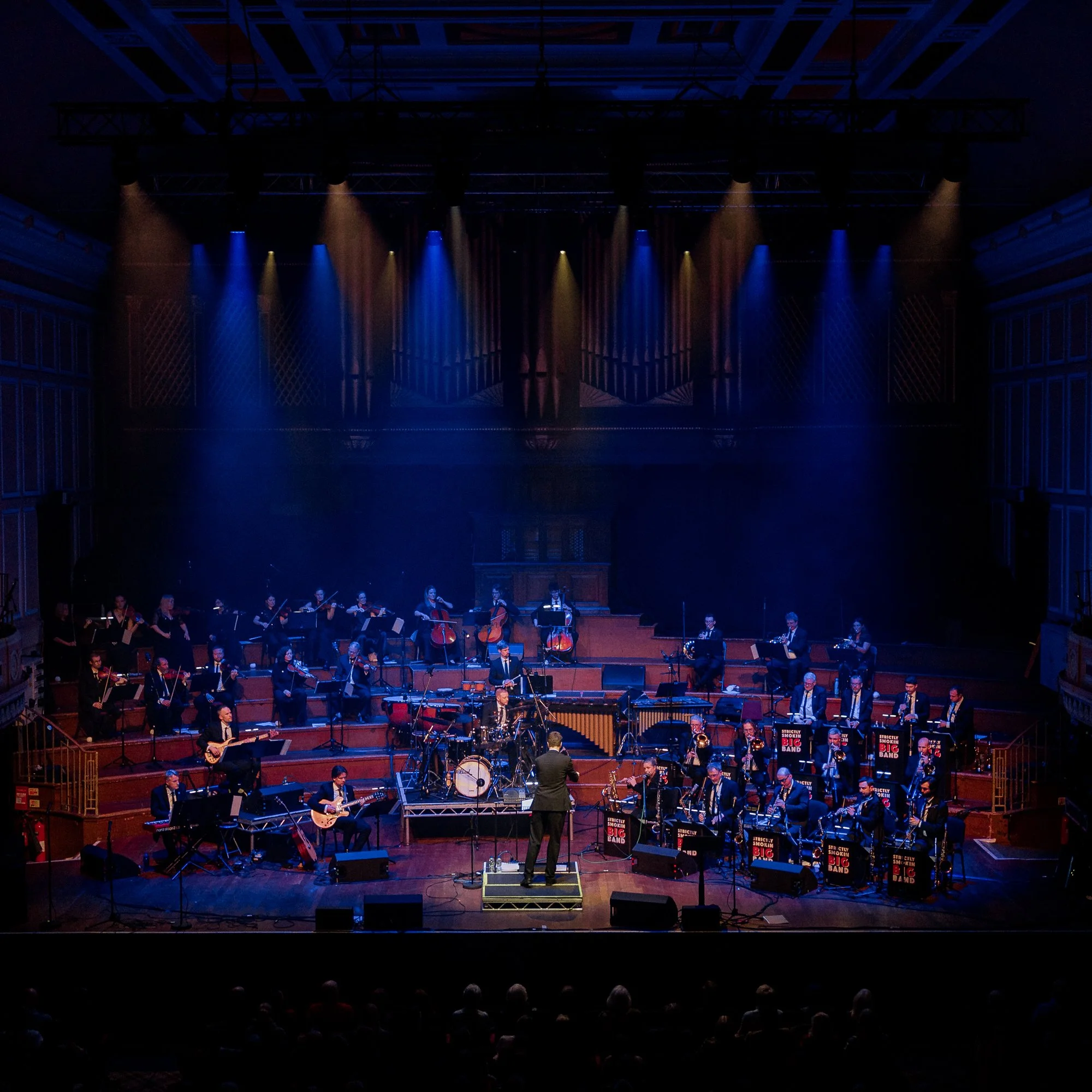A large orchestra performing on stage in a concert hall with colorful stage lighting and a conductor in the center.