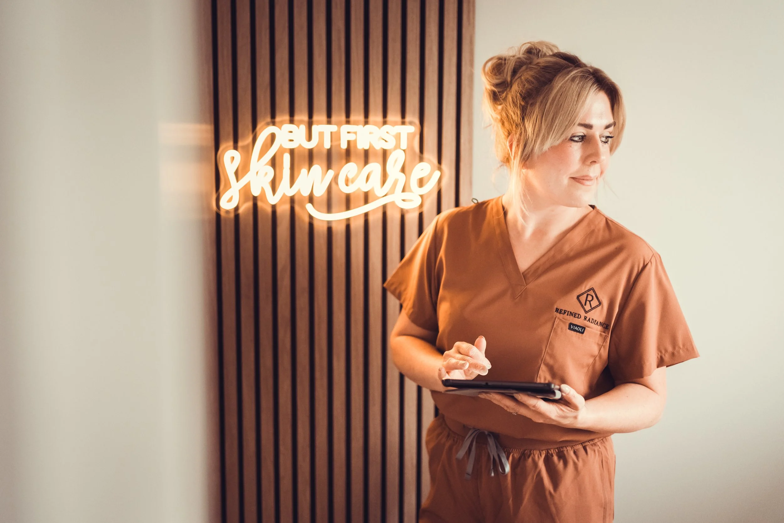 A woman in brown scrubs standing in front of a wall with a neon sign that says "But First Skin Care".