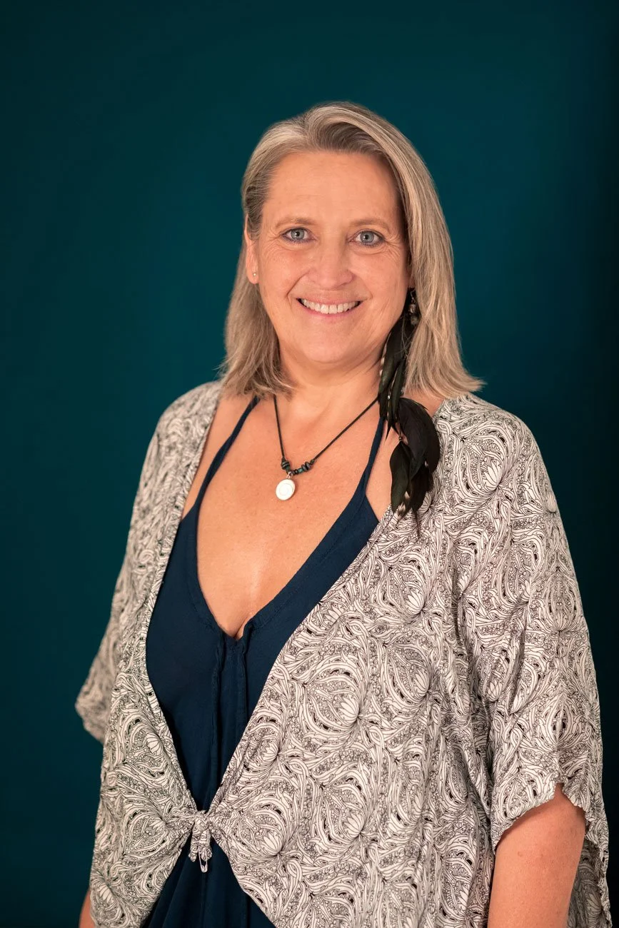 Porträt einer lächelnden Frau mit grauen Haaren, blauen Augen, einem schwarzen Kleid und einem weich fallenden, gemusterten Überwurf, vor einem dunklen Türkiston-Hintergrund.