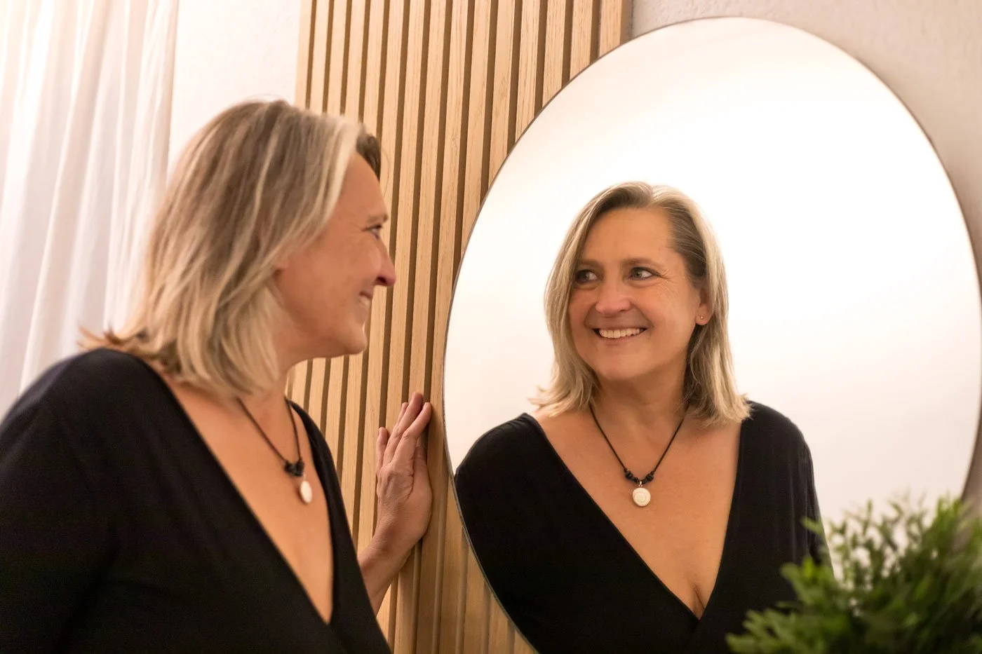 Eine Frau lächelt beim Blick in einen runden Spiegel, während sie ihn berührt. Sie trägt eine schwarze Kleidung und eine Halskette. Im Hintergrund sind vertikale Holzpaneele zu sehen.