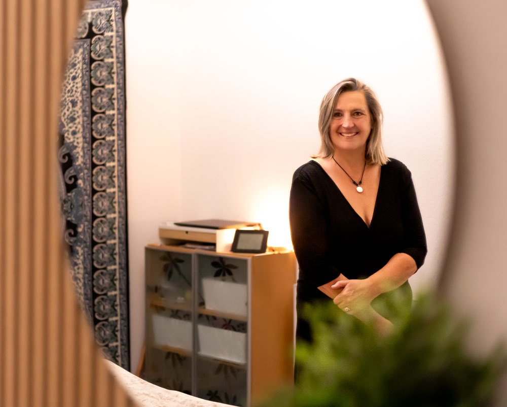 Eine lächelnde Frau steht in einem Raum, sichtbar durch eine spiegelartige Oberfläche, mit einem Sideboard und einem Bild oder Teppich mit blumigem Muster im Hintergrund.