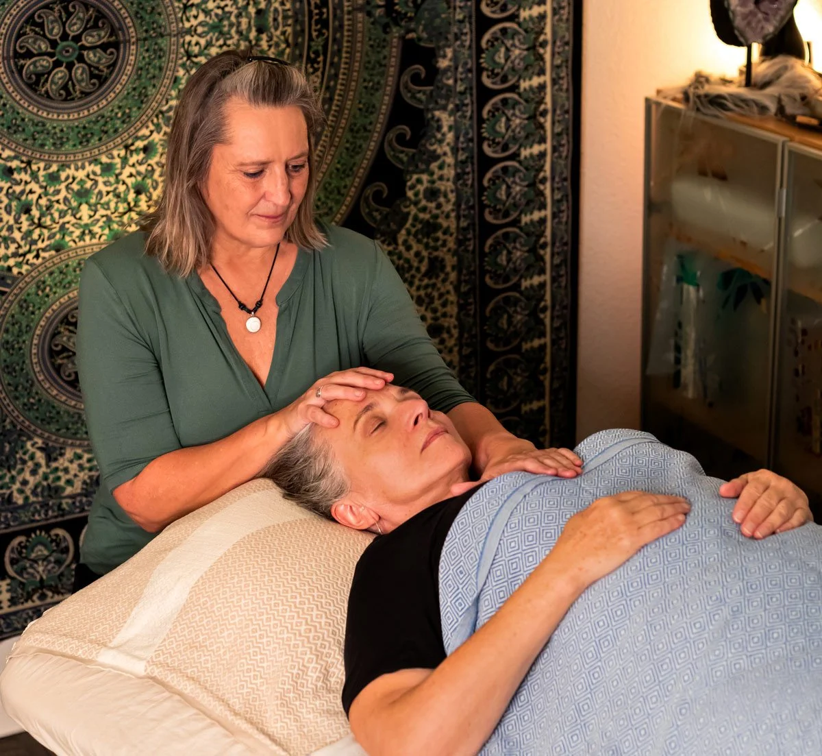 Eine ältere Frau liegt auf einer Liege und erhält eine Kopfmassage oder Heilbehandlung von einer Frau in einer grünen Bluse in einem Raum mit einer dekorativen Wand hinter ihr.