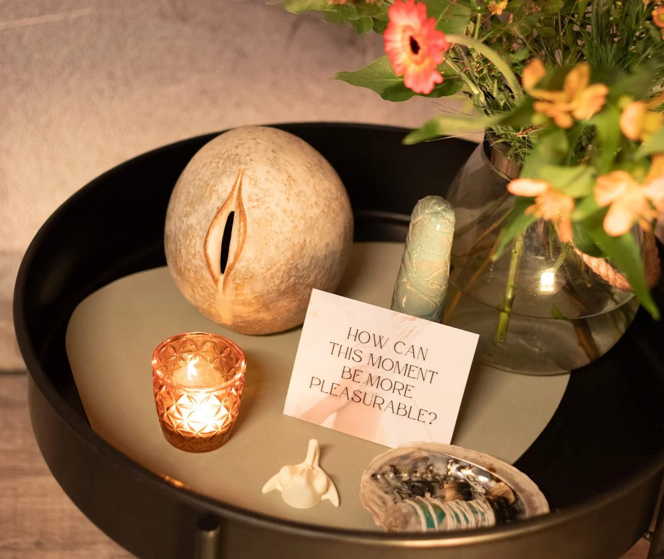 Ein schwarzer Tisch mit Dekorationsgegenständen, darunter eine Muschel, eine kleine Kerze in einem Kristallglas, ein Zettel mit dem Text 'How can this moment be more pleasurable?', eine dekorierte Vase mit Blumen, ein ovales Artefakt und eine kleine weiße Skulptur, die zusammen eine ruhige, meditierende Atmosphäre schaffen.