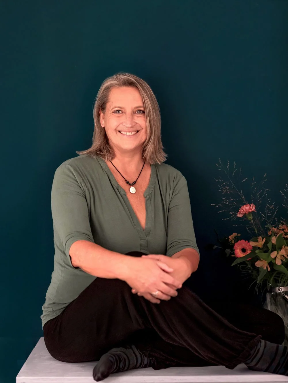 Eine lachende Frau mit Schulterlange Haare in einem grünen Oberteil sitzt gegen eine dunkelgrüne Wand, rechts eine Vase mit bunten Blumen.