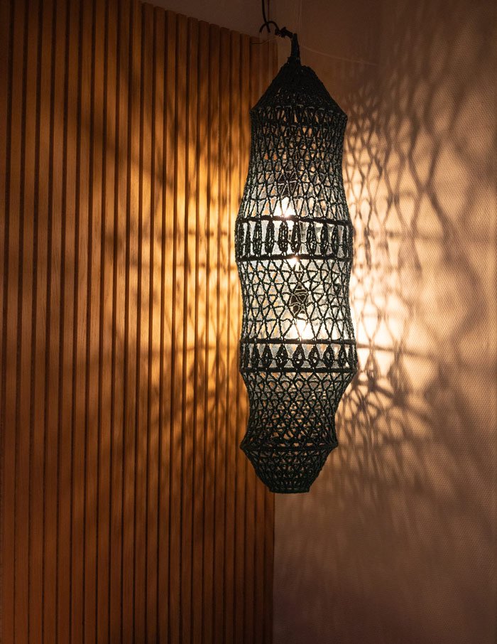 Hängeleuchte aus schwarzem, geflochtenem Material wirft komplexe Schattenspiele an die Wand, neben einer vertikalen Holzpaneelwand.