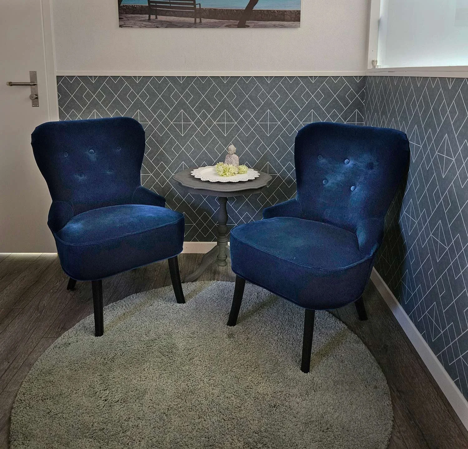 Zwei blaue Sessel vor einer kleinen, schwarzen Beistelltisch, auf dem eine Deko mit einer kleinen Statue und weißen Blumen liegt, in einem Raum mit geometrisch gemusterter Tapete und einem geraden Wandbild.
