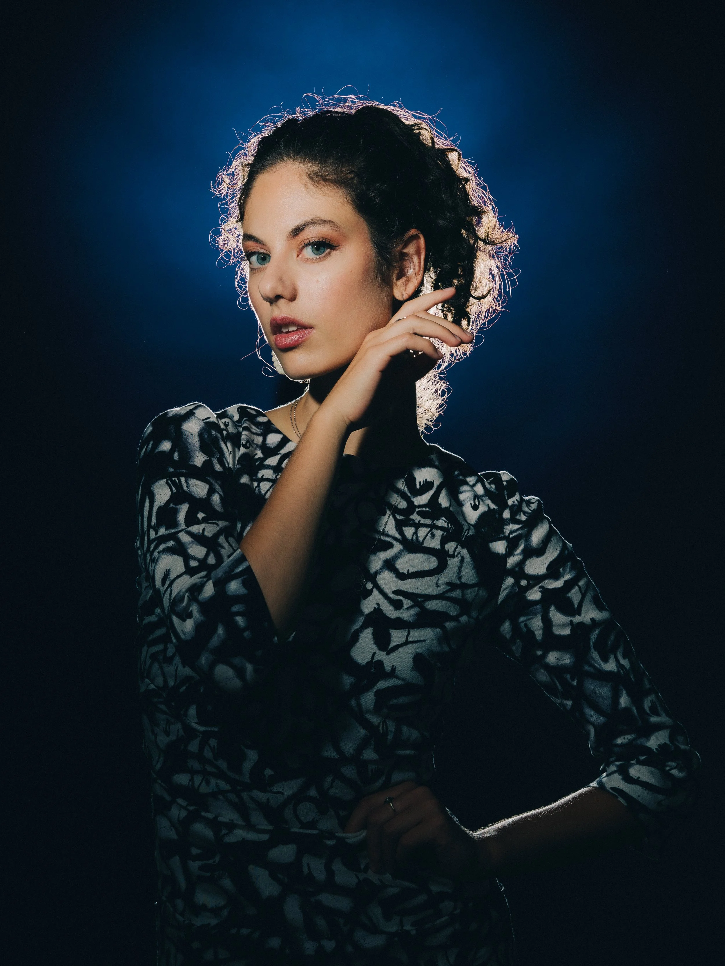 A portrait of a young woman with curly hair, wearing a patterned black and white top, standing against a dark background with a blue light behind her. She has piercing blue eyes and is looking at the camera with her hand near her face.