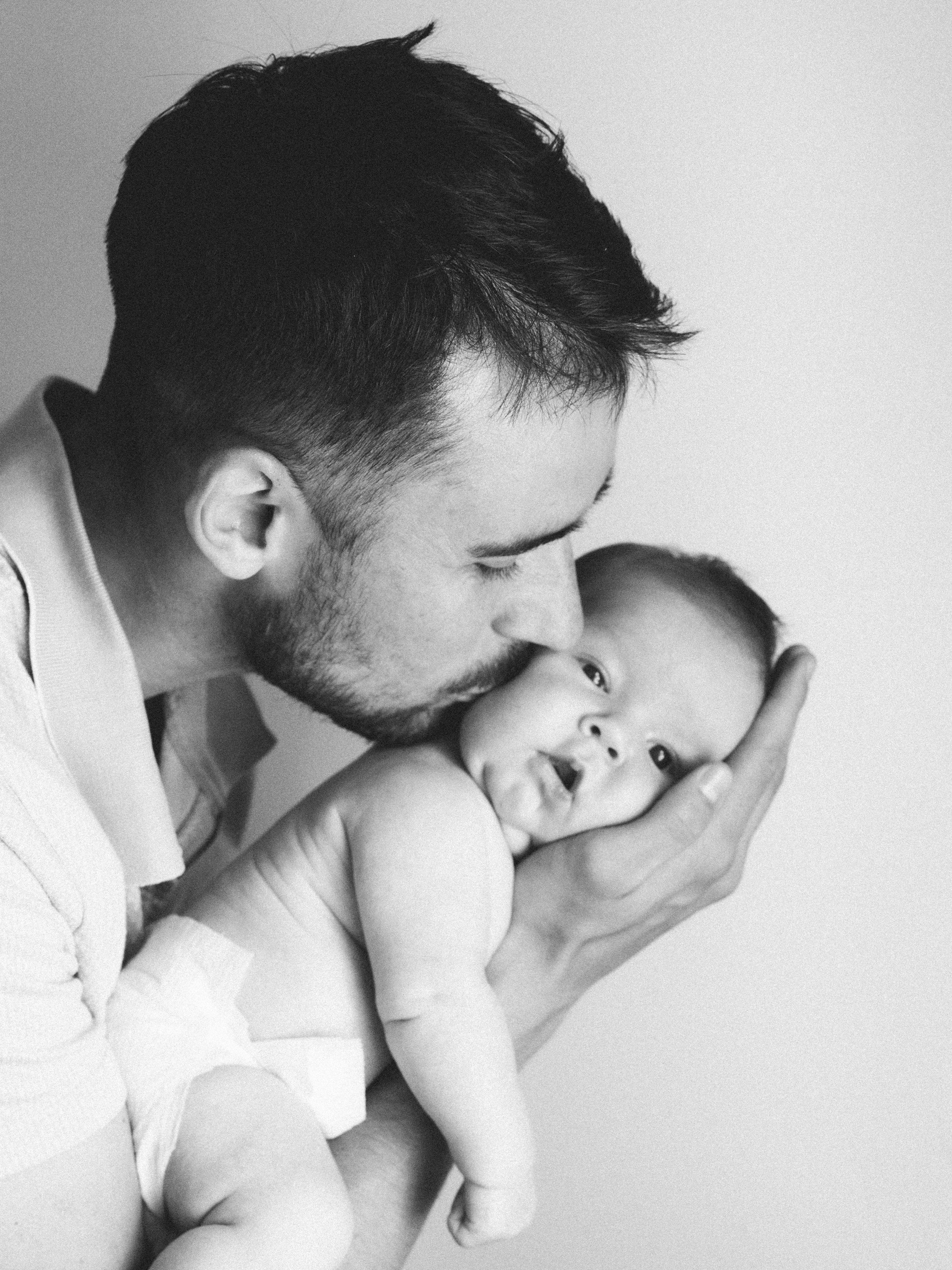 A black and white photo of a man gently kissing a baby on the forehead, with the baby looking directly at the camera.