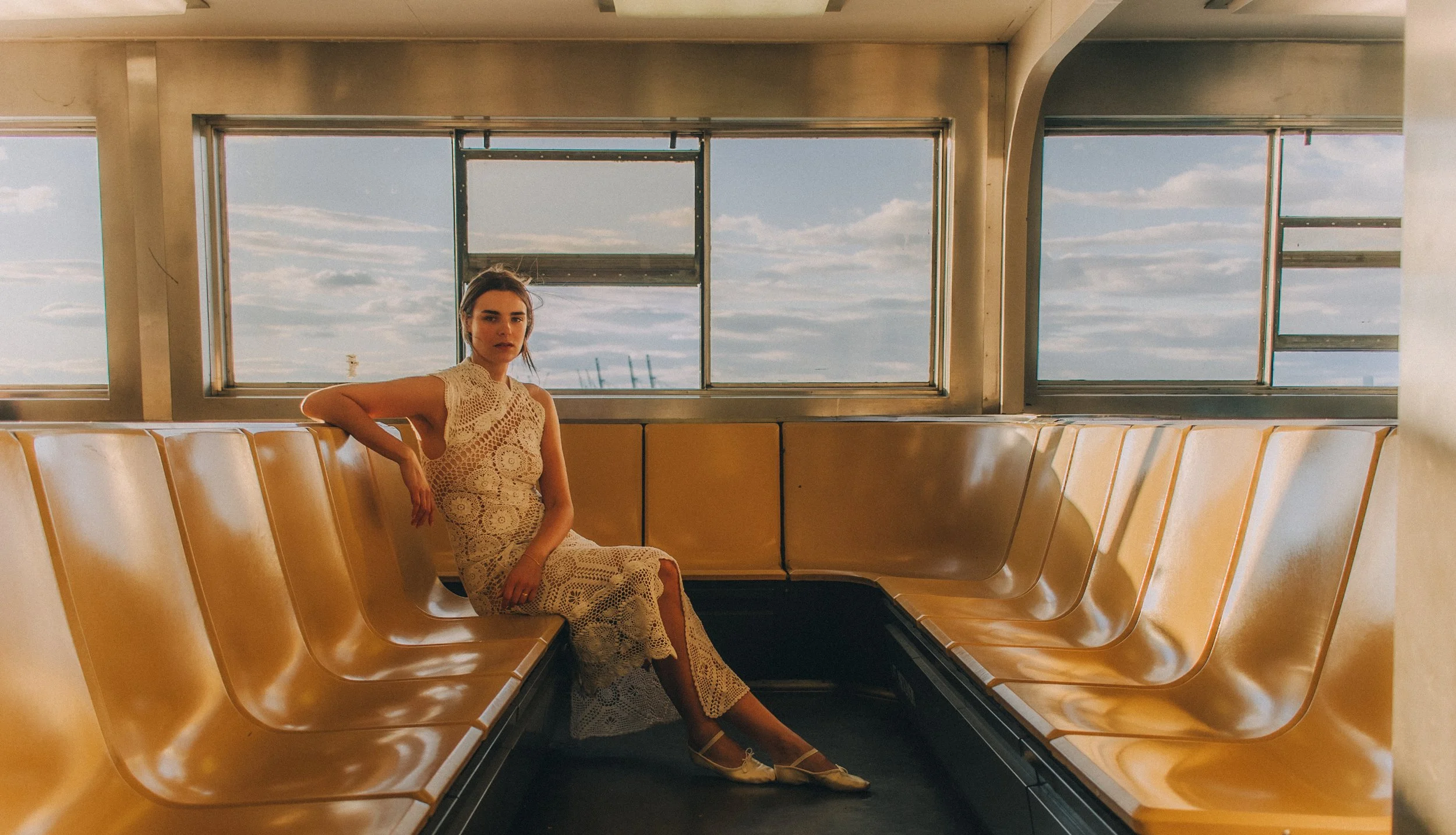 A woman in a cream-colored crochet dress sitting on a row of yellow seats inside a bus or train with large windows showing a cloudy sky outside.