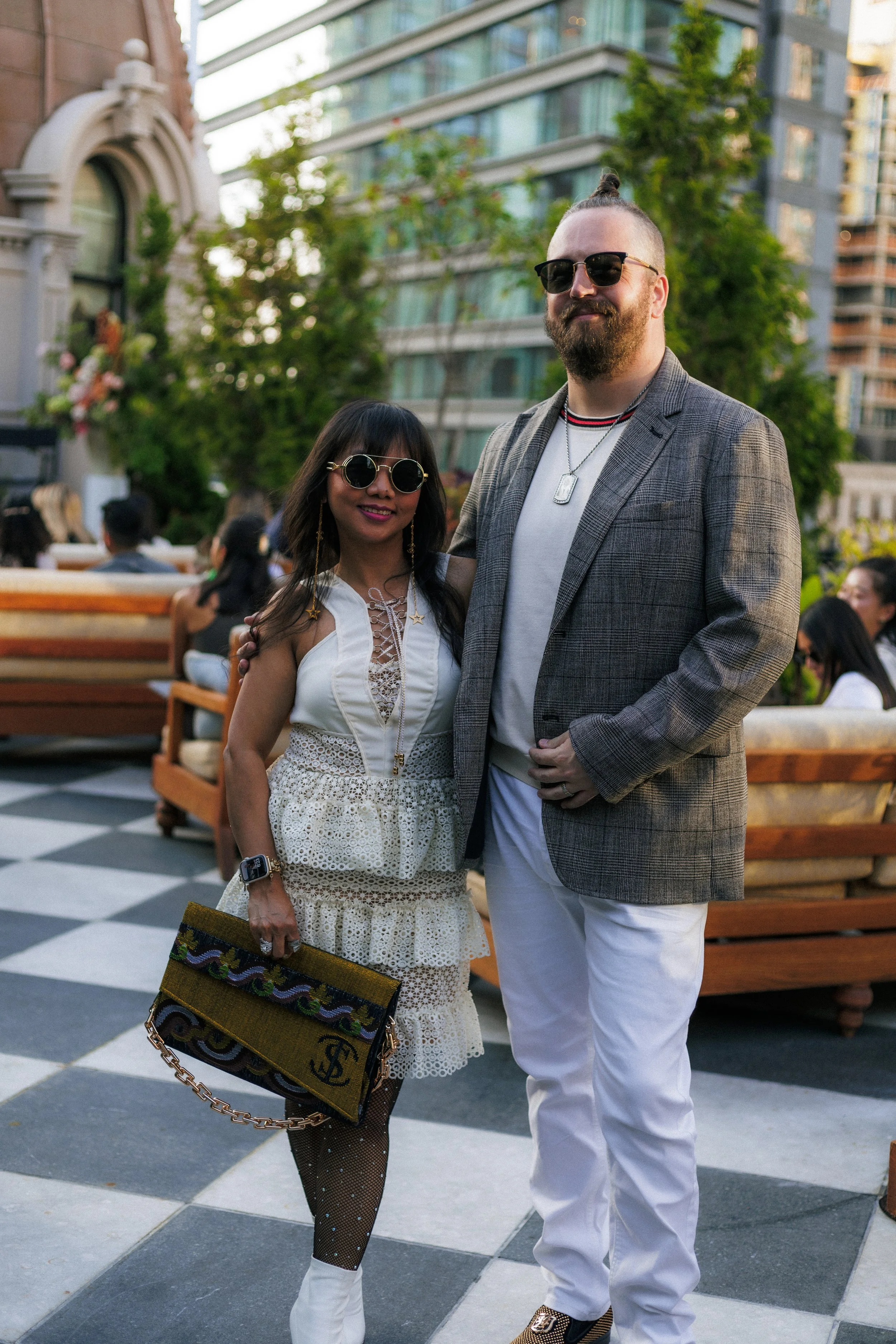 A man and a woman are posing together outdoors in a city setting. The woman is wearing a white dress with lace detailing, black fishnet stockings with polka dots, white boots, and sunglasses. She is holding a patterned designer handbag. The man is dr