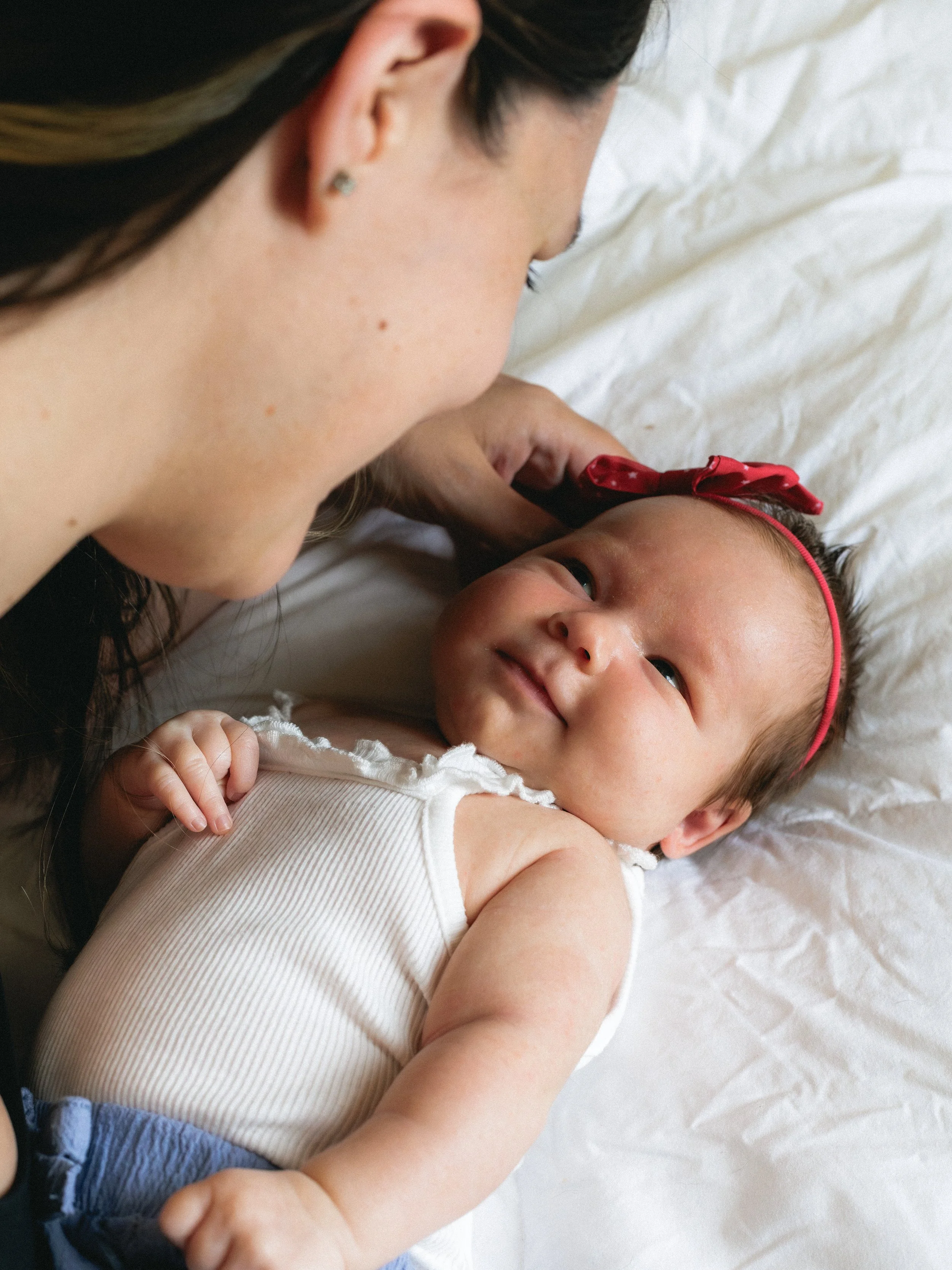 A young woman with dark hair and earrings leans over and gently kisses a baby girl lying on a white bed, who is looking up with a smile and a red headband.