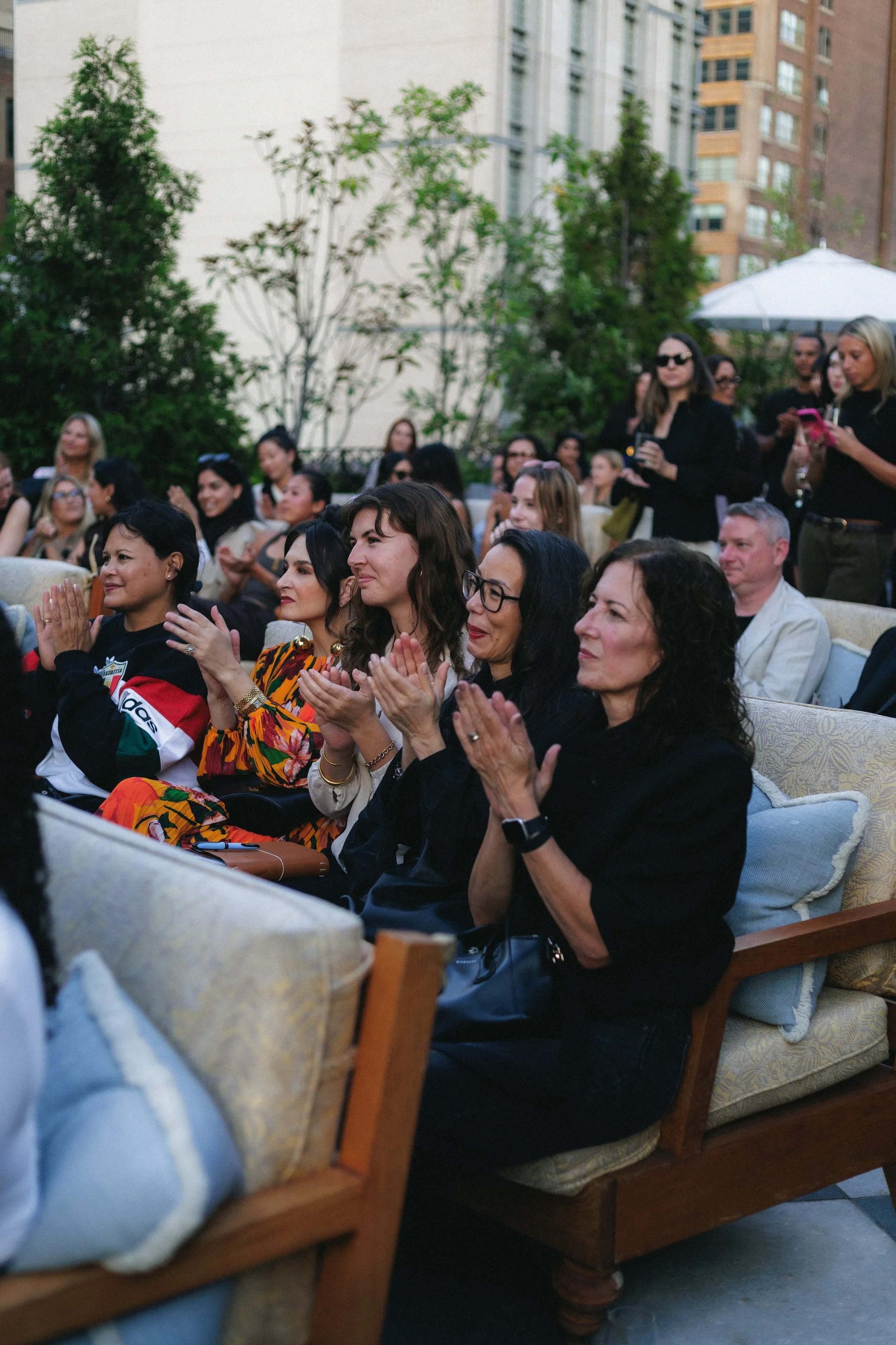 An outdoor event with a seated audience of diverse women and men, some clapping and others using their phones, in front of a background of trees and city buildings.