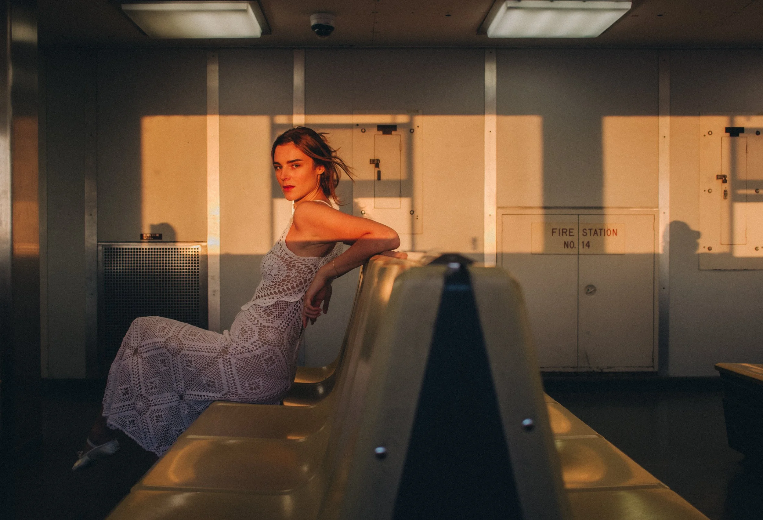 A woman with light brown hair wearing a white crocheted dress sitting on a yellow airport bench, with the sunset casting warm light and shadows in the background.