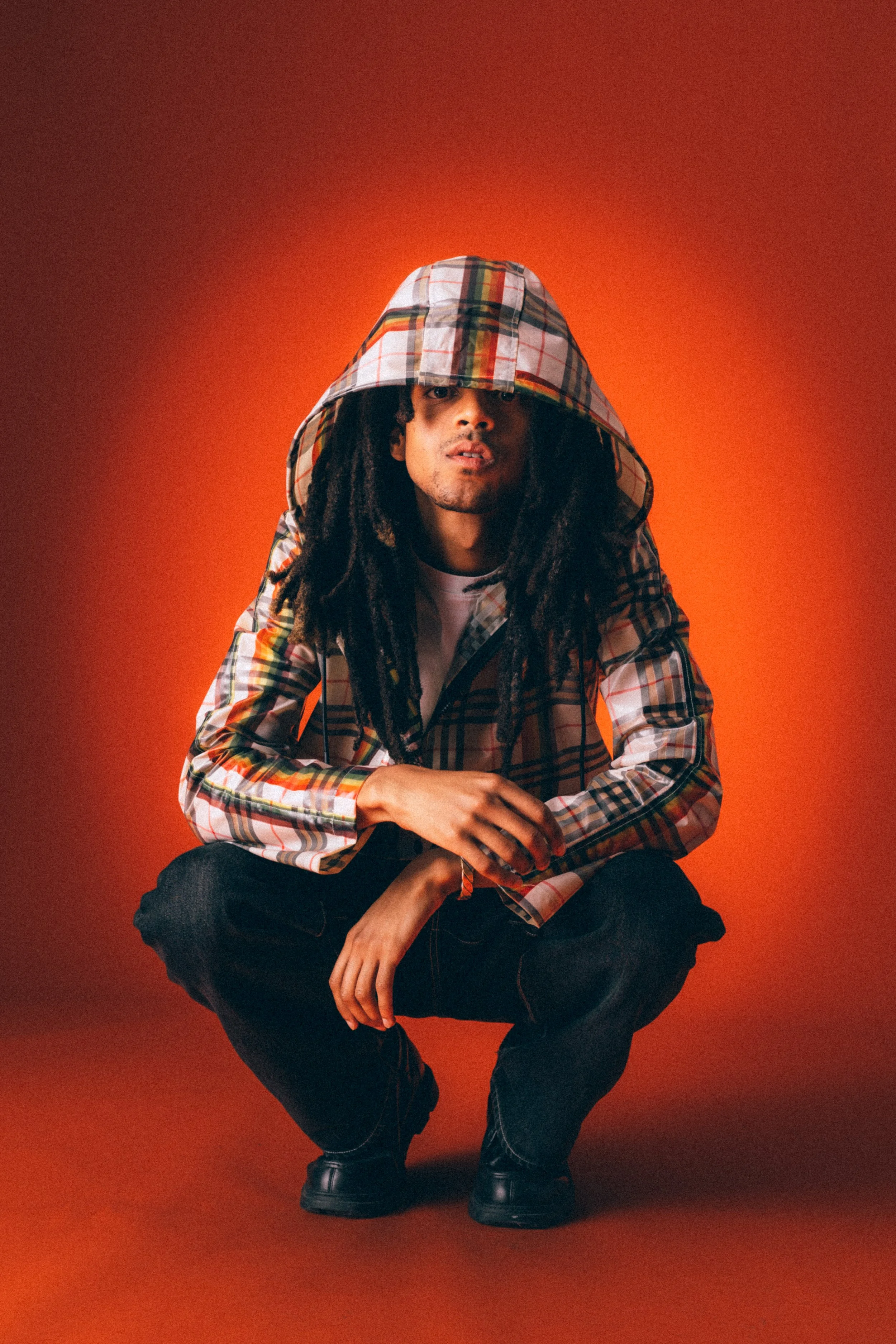 A person with dreadlocks crouching against an orange background, wearing a hooded plaid jacket, black pants, and black shoes.