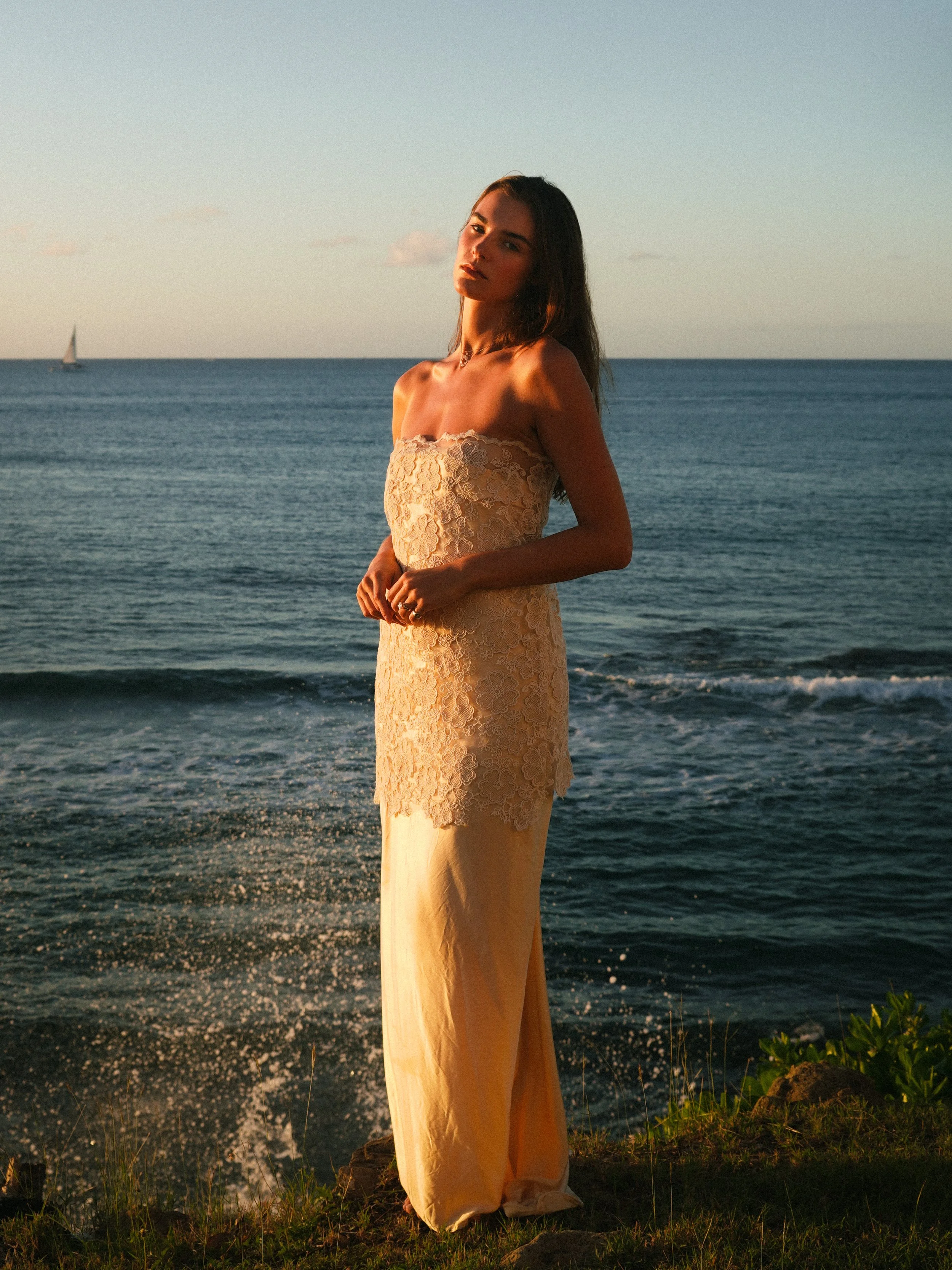 A woman in a strapless lace dress standing on a grassy area near the shoreline at sunset, with the ocean and a sailboat in the background.