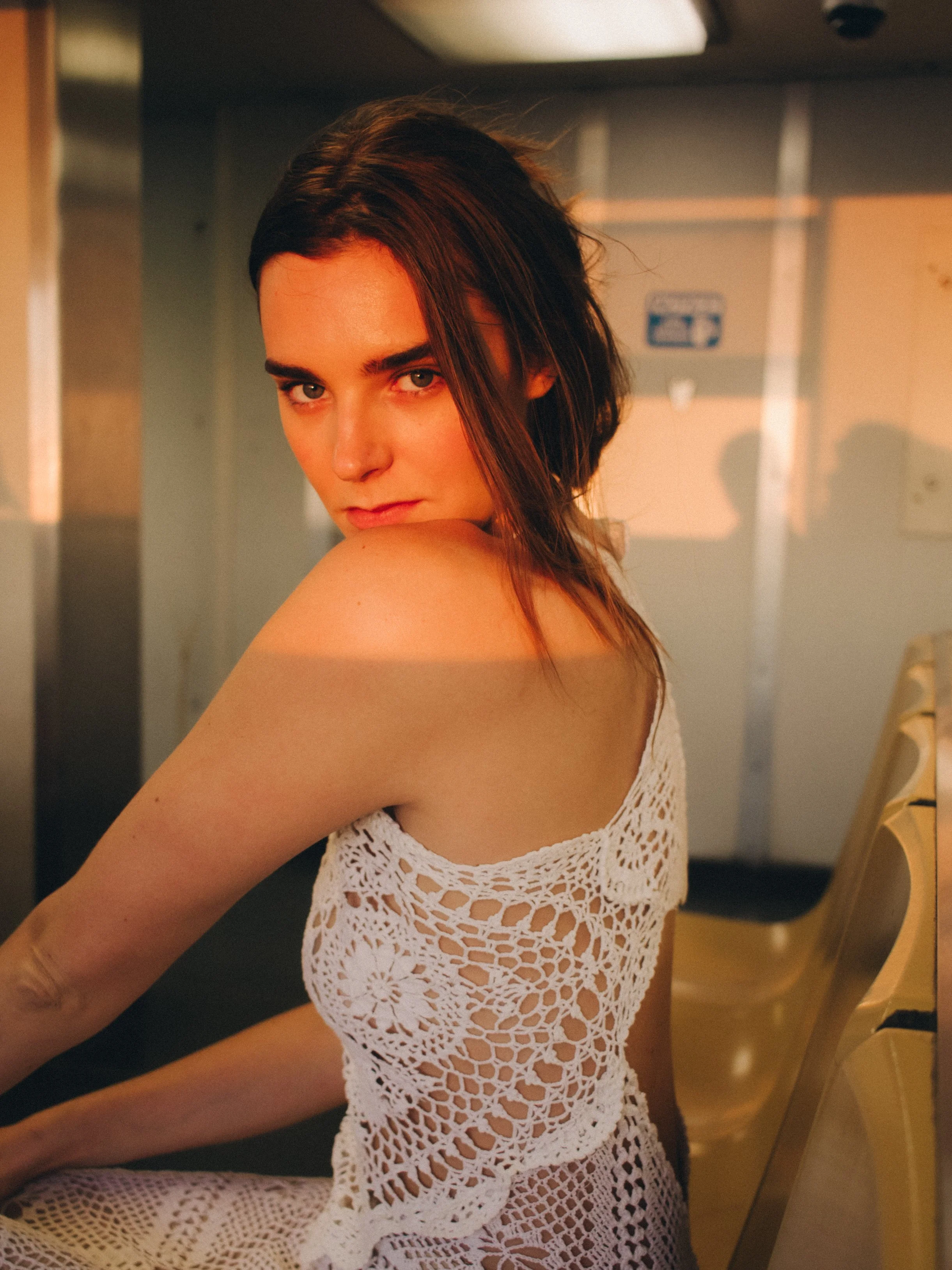 A young woman with short brown hair and light skin wearing a white crochet dress sitting at a table in a room with yellow chairs and beige walls.