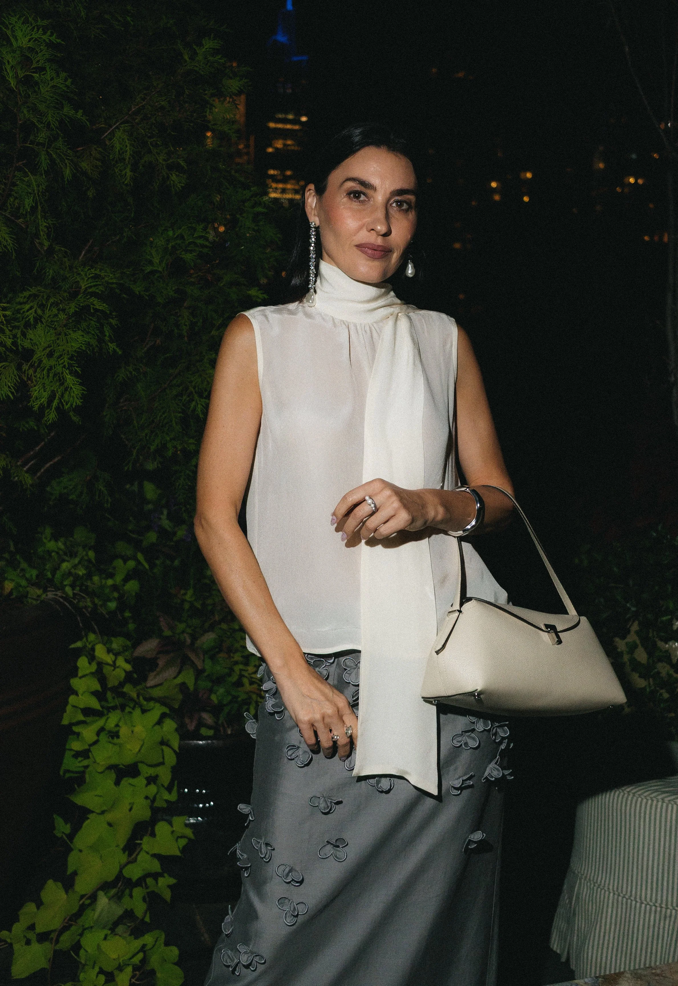A woman dressed in a sleeveless white top and gray skirt with butterfly patterns holds a cream handbag. She has dark hair, wearing long earrings, and a ring on her finger. The background is dark with some city lights and green foliage.