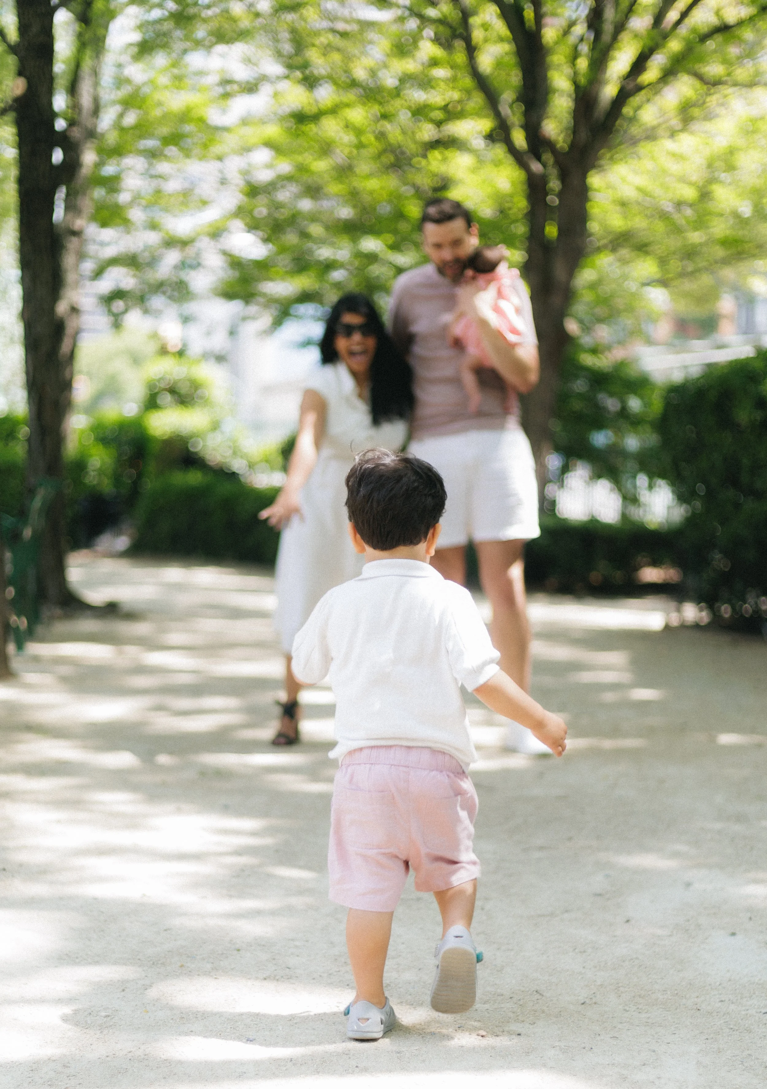 A young boy running in a park, facing away from the camera, with two adults and a small girl in the background. The adults and girl are smiling and walking among trees under a sunny sky.