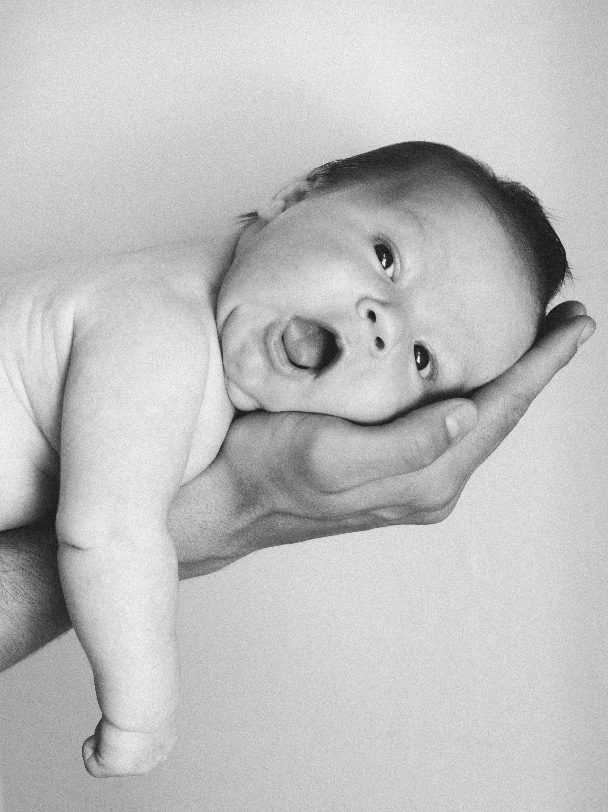 Black and white photograph of a newborn baby being gently held in an adult's hand, the baby has an open mouth and wide eyes.