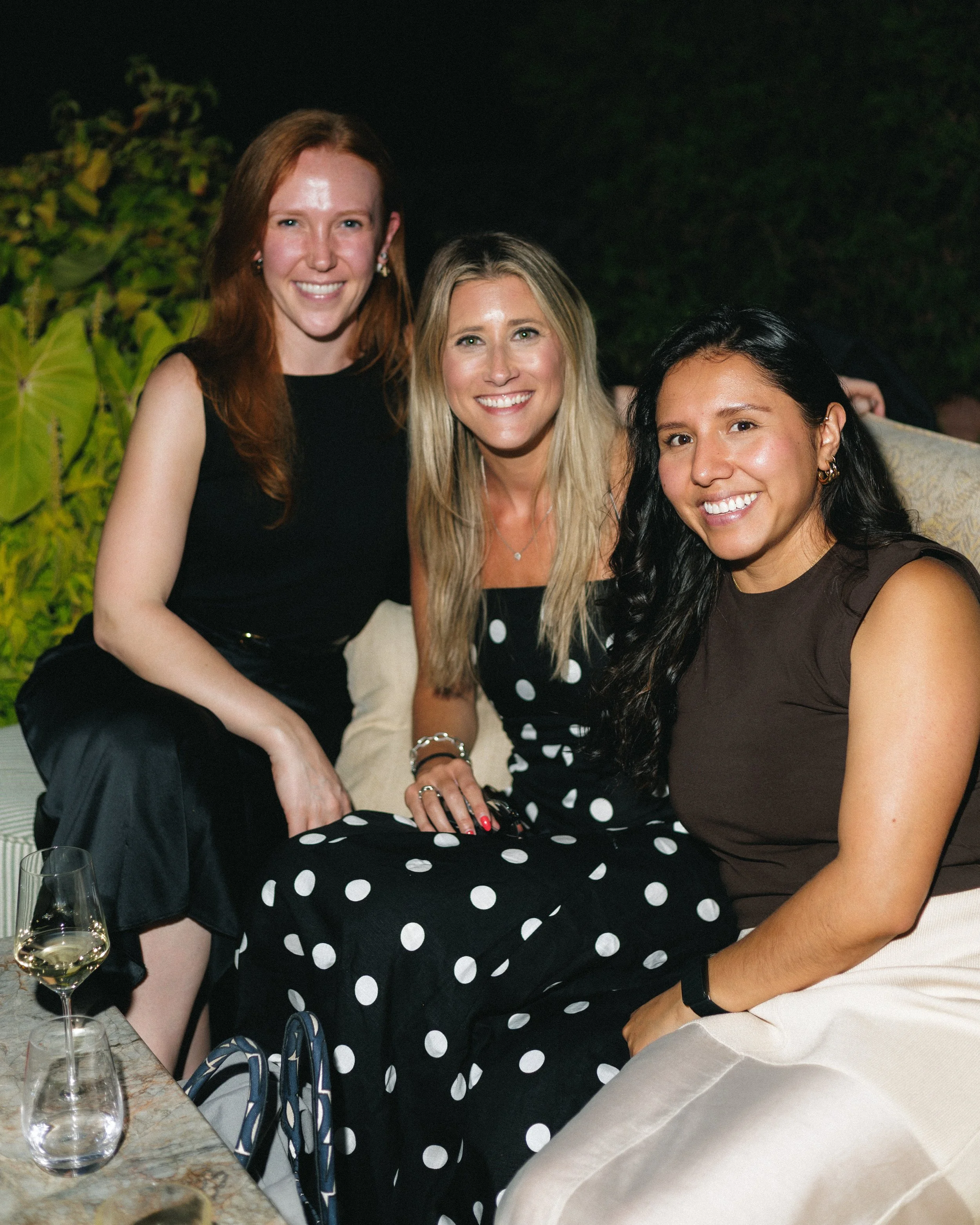 Three women sitting together at a dinner party, smiling for a photo, with wine glasses on the table and a lush green background.