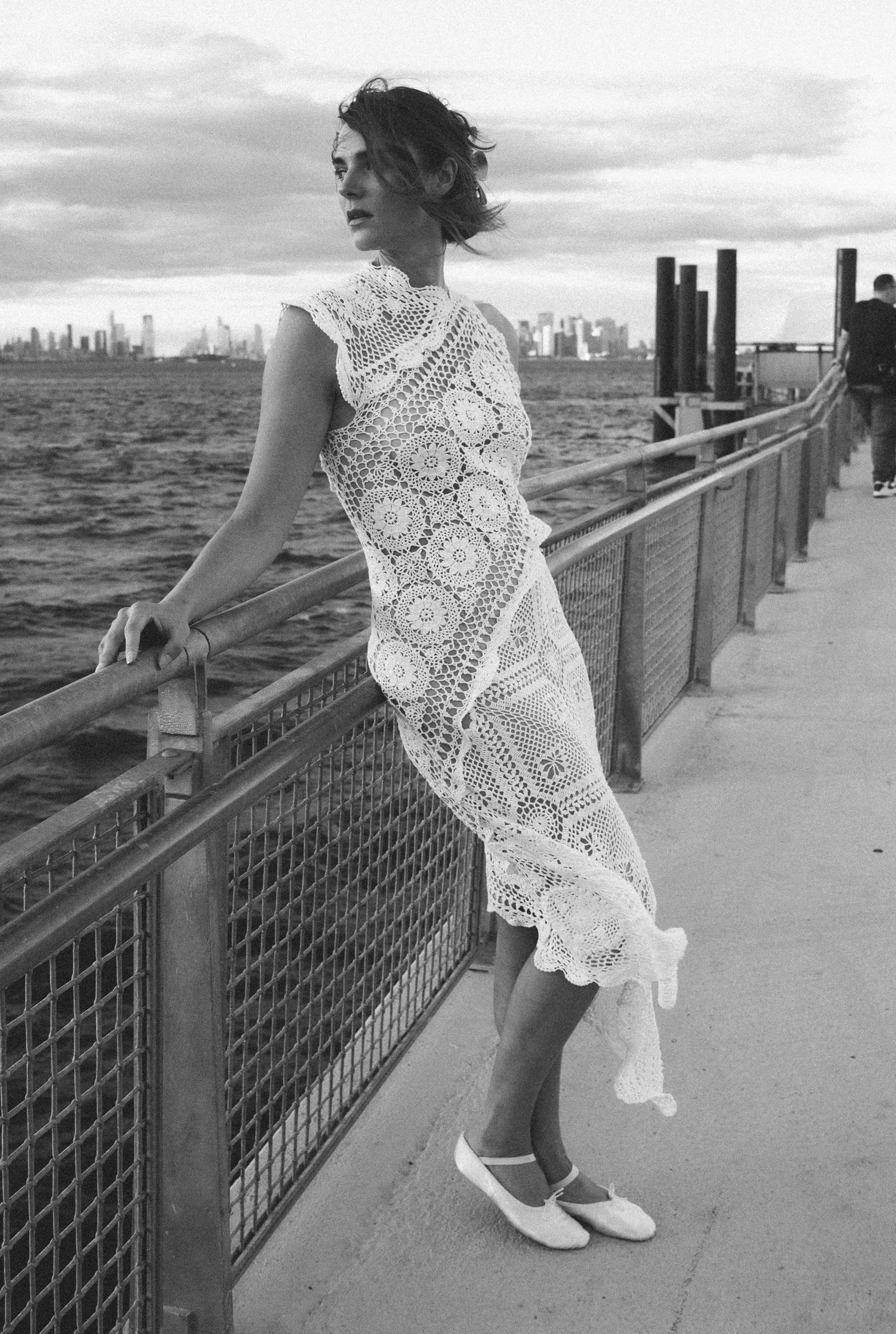 A woman in a lace dress and ballet shoes standing on a pier by the water with a city skyline in the background, black and white photo