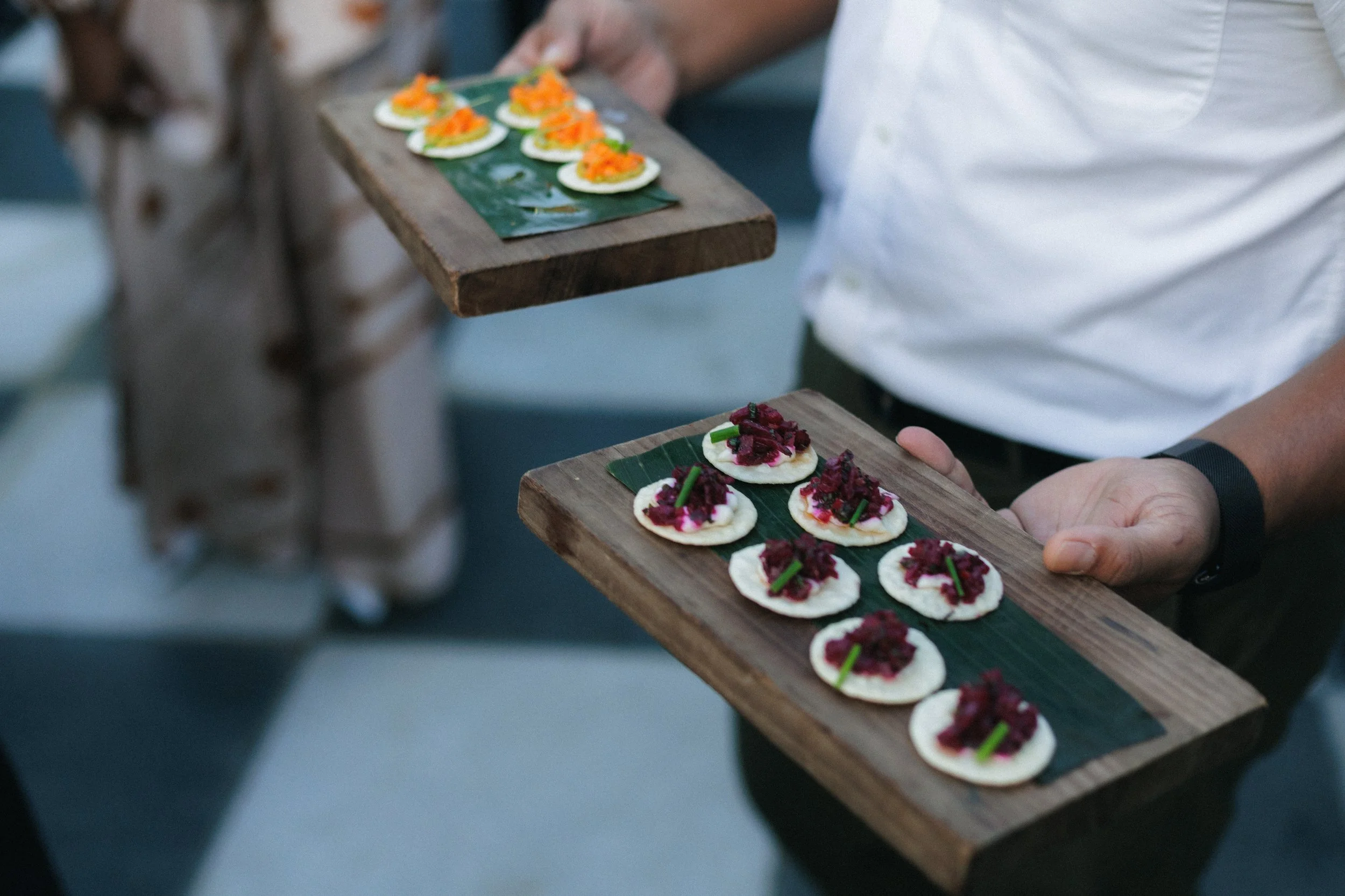 Person holding two wooden platters with small canapés, topped with colorful ingredients, possibly for an appetizer or snack.