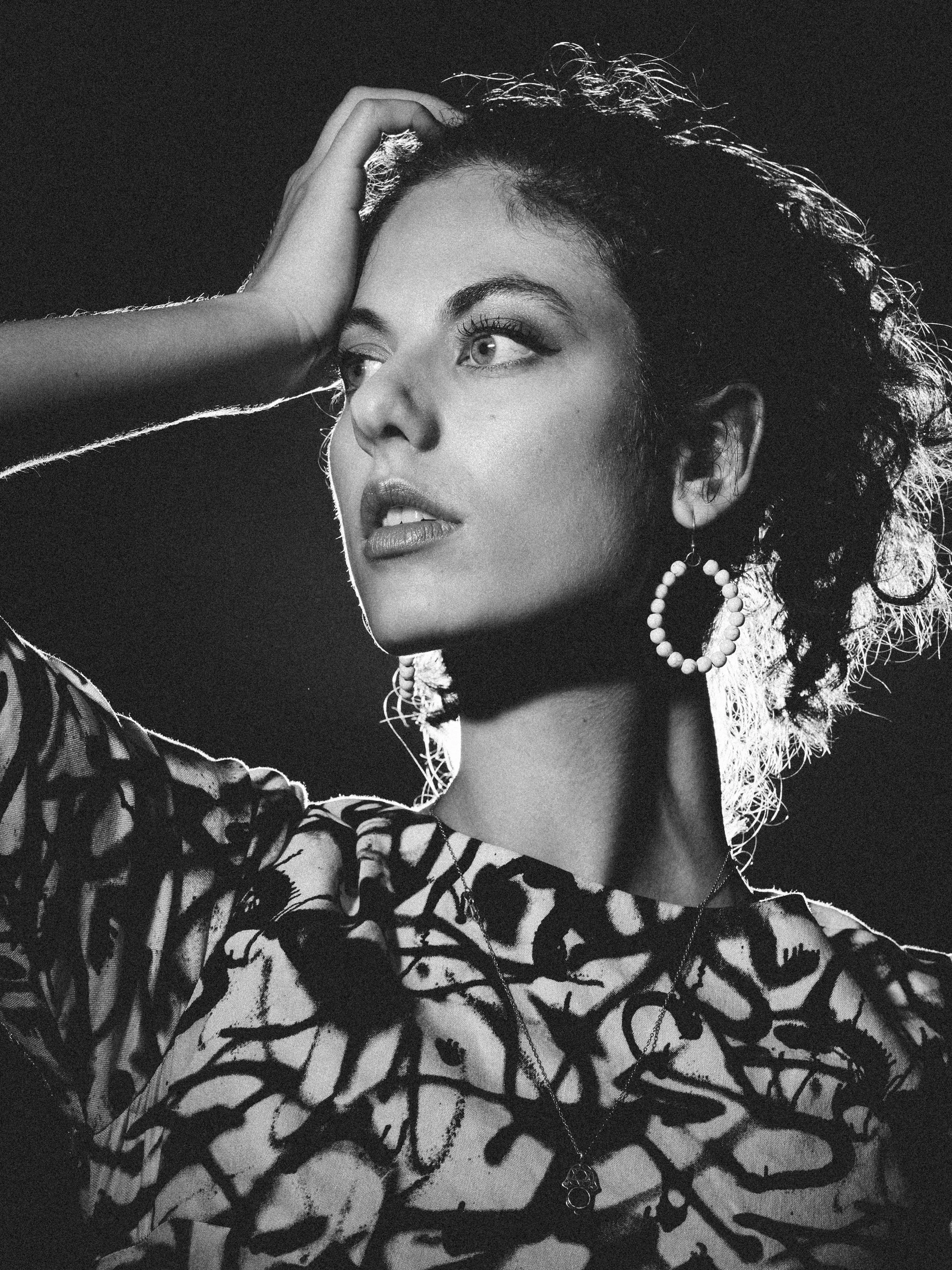 Black and white portrait of a woman with curly hair, wearing large hoop earrings, a patterned top, and a necklace, touching her forehead with one hand.