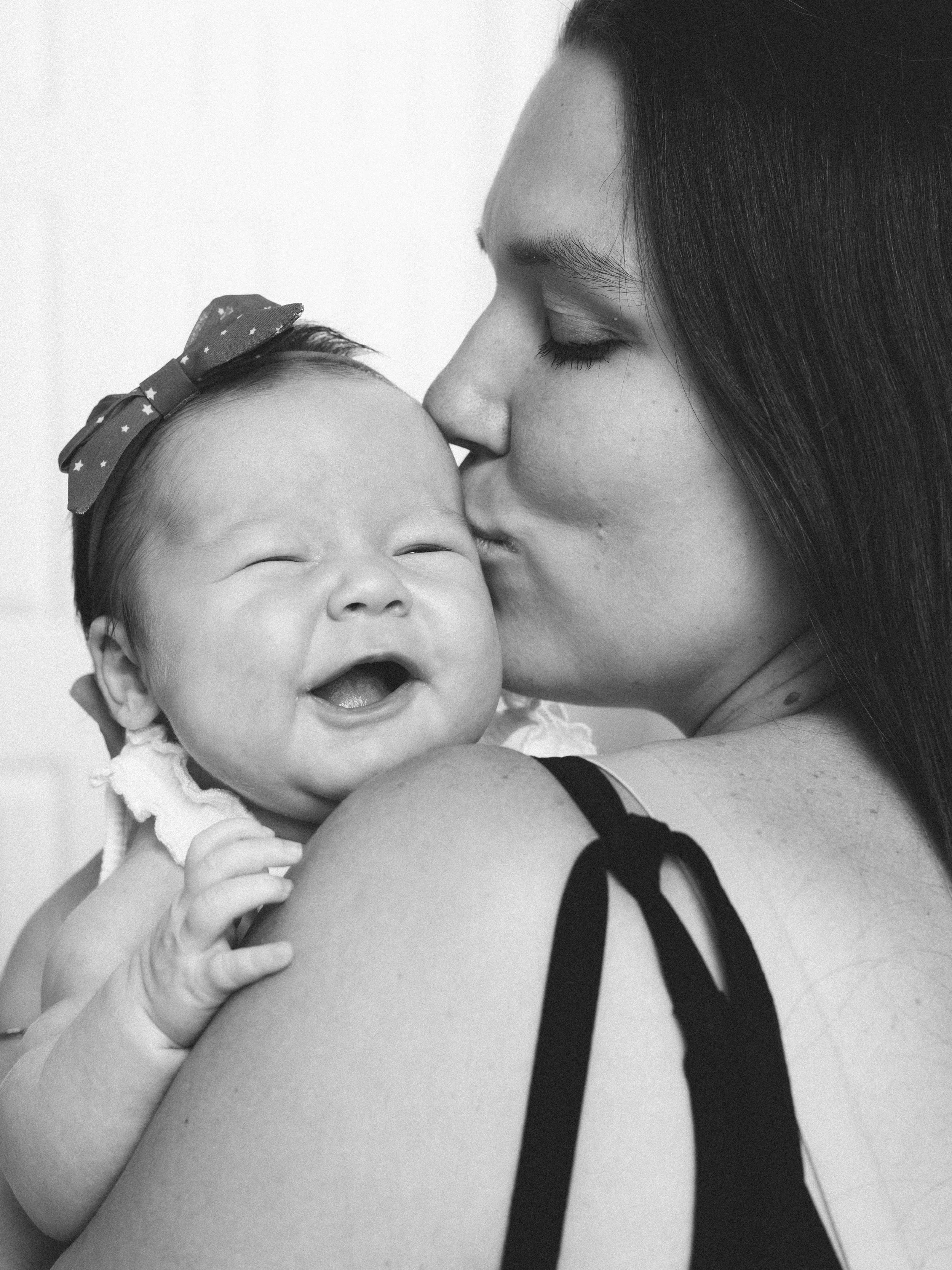 A woman with long dark hair gently kisses a smiling baby girl with a bow in her hair, in a close-up black-and-white photograph.